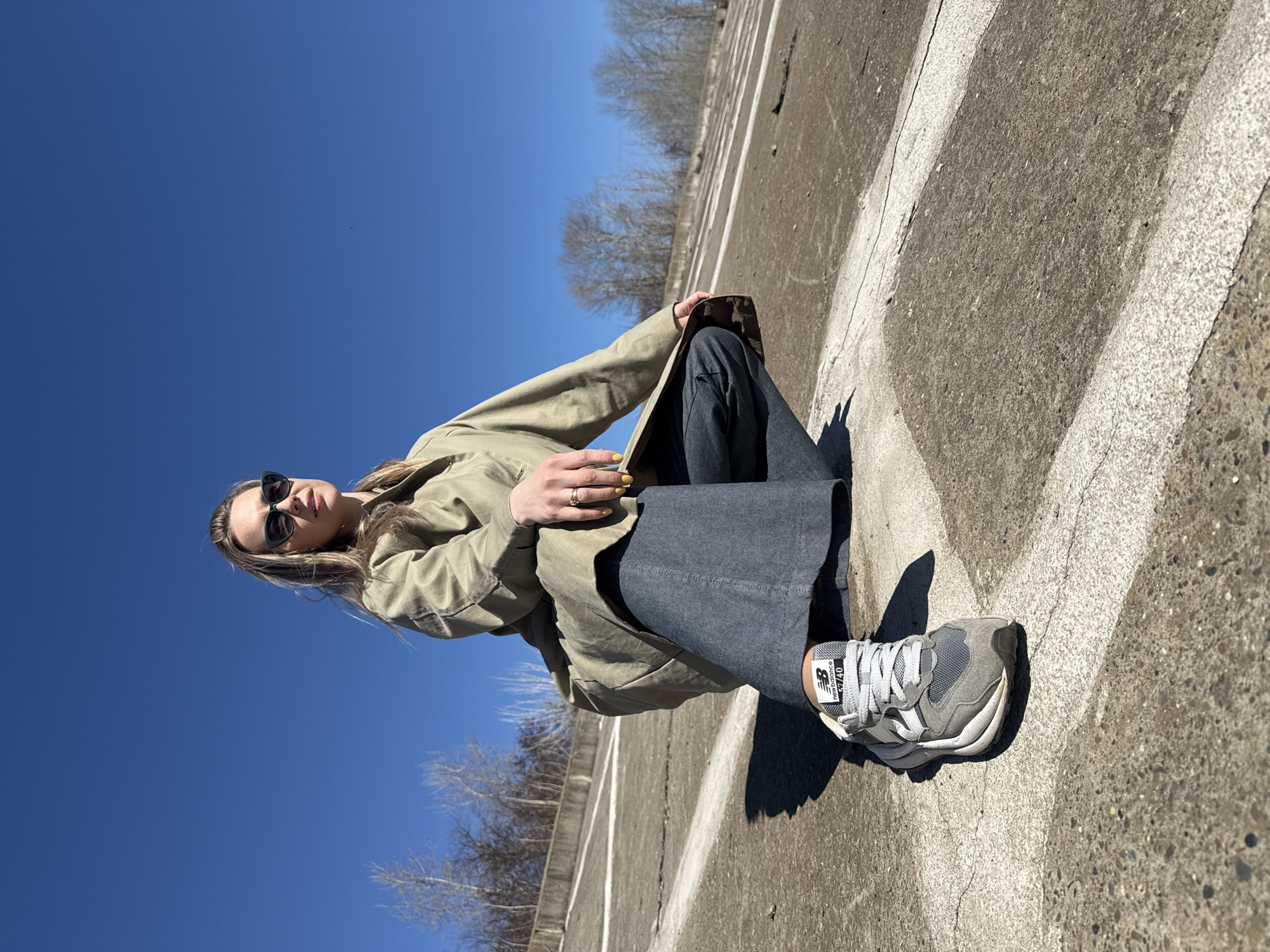 A woman is sitting on a paved surface with white lines, likely a parking lot or similar area, under a clear blue sky. Bare trees are visible in the background. She is wearing sunglasses, a khaki-colored trench coat, and grey pants. Her right leg is extended with a grey and white New Balance sneaker. She has a yellow manicure. The image has been rotated clockwise. The location is assumed to be in Chaikovsky, Russia.