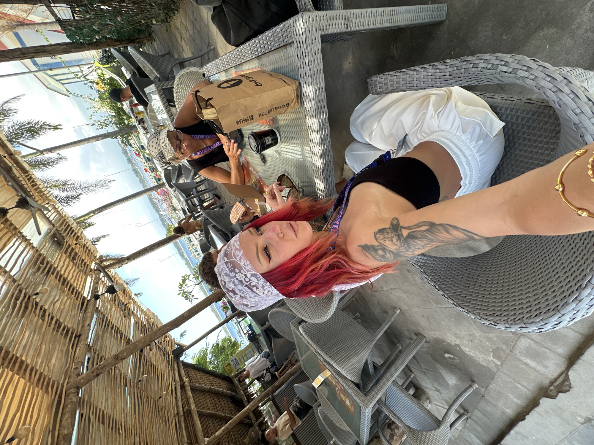 This photograph captures a scene in an outdoor dining area, likely in Unknown, Indonesia, indicated by the presence of tropical foliage and the woven wooden structure on the left. A woman with red hair, wearing a white lace head covering and a black top, is taking a selfie. She has a visible tattoo on her arm and is wearing a gold bracelet. Beside her, another person sits at a woven gray table. A brown paper bag with the text "69SLAMOFFICIAL" is on the table, along with a black cylindrical container. Other people can be seen in the background, seated at similar tables. In the distance, there's a view of a body of water, possibly a bay, with buildings and boats. The setting suggests a relaxed, casual atmosphere.