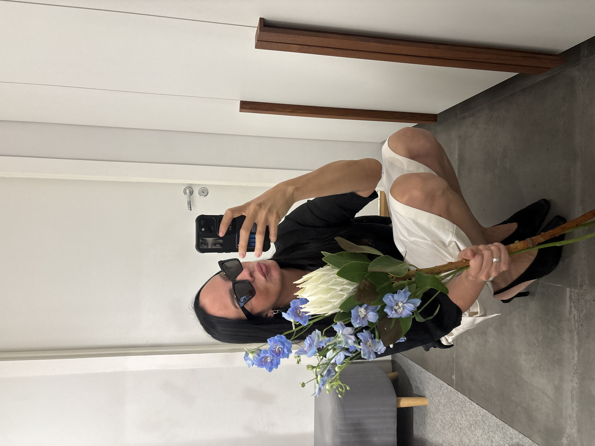 The image shows a woman squatting and taking a selfie in what appears to be a modern indoor space, possibly a home or dressing room. She is holding a bouquet of flowers, including blue delphiniums and a large white protea. The woman is wearing black sunglasses, a black top, and white shorts or a skirt. She has long dark hair and is wearing black high-heeled shoes. The background includes a white door with a silver handle on the left and white closet doors with vertical wooden accents on the right. There is a gray upholstered bench with wooden legs visible on the far left. The floor is made of large gray tiles. No specific location within Chaikovsky, Russia is identifiable from the image.