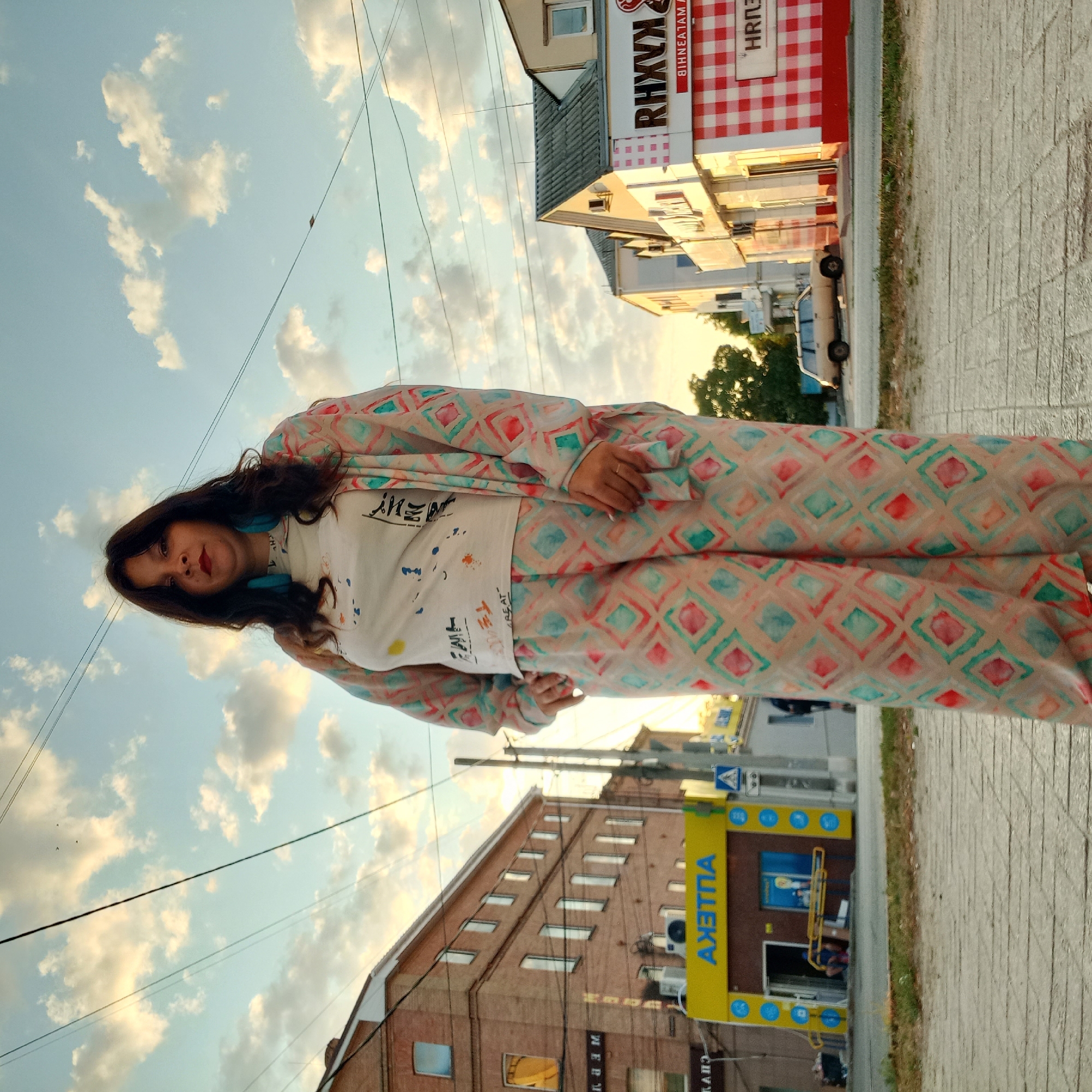 A full-shot, eye-level, outdoor medium photograph captures a woman standing on a tiled sidewalk. She has dark hair, red lipstick, and is wearing teal-colored earrings. Her outfit consists of a white t-shirt with a colorful abstract print and loose-fitting pants and a matching long-sleeved open cardigan adorned with a diamond pattern in shades of pink, teal, and beige.
The background reveals a street scene with buildings, power lines, and a cloudy sky illuminated by the golden light of sunset. To the left, a brick building with multiple windows is visible, and a yellow storefront with blue text and circular motifs is present. Further to the right, another building with a sign reading "RHXVK" is seen, featuring a red and white checkered pattern. A light-colored station wagon is parked on the street. The overall setting appears to be an urban environment. This location is the intersection of Volodymyra Vernadskoho Street and Oleksiy Vasyliev Street in Kropyvnytskyi, Ukraine.