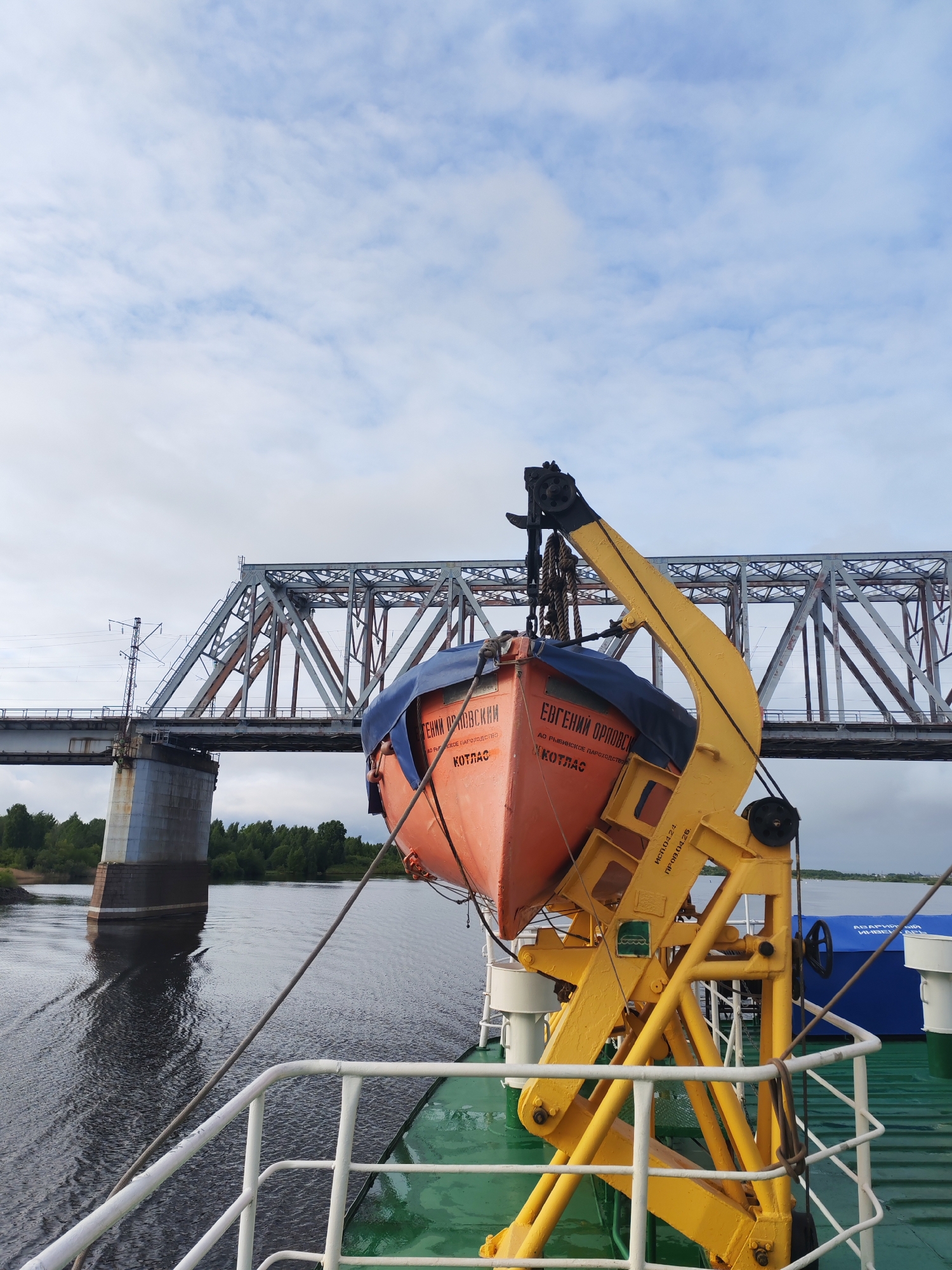 The image depicts a lifeboat hanging from a yellow crane on a ship. The lifeboat is orange and has the words "Евгений Орловский" and "Котлас" written on it in Cyrillic script. A blue tarp partially covers the boat. In the background is a steel truss bridge over a body of water, with trees visible on the far bank. The sky is overcast with white and gray clouds. The ship's railing is visible in the foreground.
Based on the OCR text mentioning "Котлас", the bridge likely spans the Northern Dvina River near the city of Kotlas, though more specifically, the photo location is the city of Нифантово, Russia, due to the presence of the Nifantovo bridge.