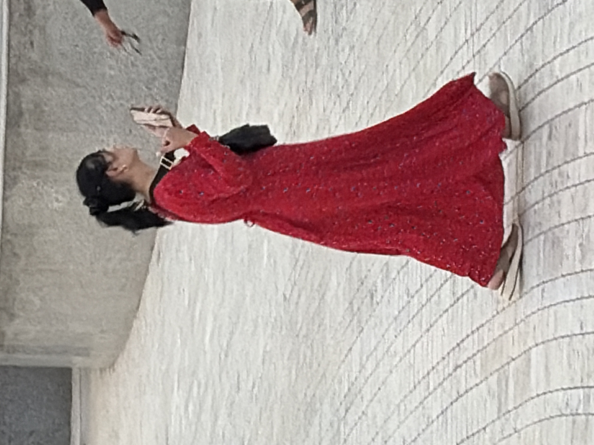 A woman wearing a long, red, floral-patterned dress and a black purse slung over her shoulder, is walking to the right, holding a phone in her right hand. She has dark hair pulled back in a ponytail and is wearing glasses. Her feet are clad in slip-on sandals with white soles. The ground is paved with light-colored, rectangular tiles arranged in a curved pattern. In the background, a textured, light-gray wall curves to the left. Another person's arm and hand are visible on the right, holding a pair of dark-rimmed glasses.
