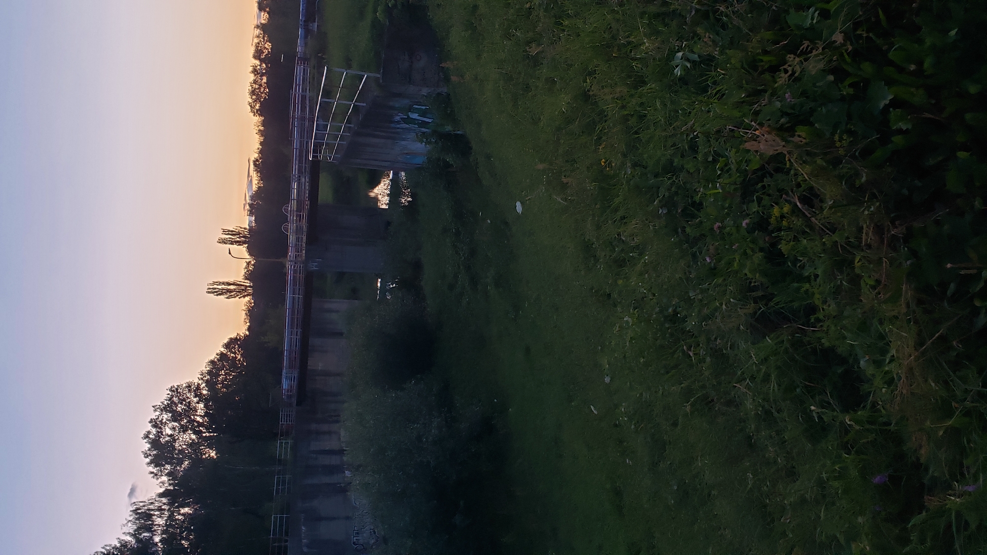 The image depicts a wide shot of a concrete bridge structure over a body of water, likely a canal or a small river, at dusk. The sky is a gradient of purple and orange, indicating sunset. Trees line the horizon behind the bridge, and the foreground is covered in lush green grass and foliage. The bridge itself has metal railings and appears to be elevated. There are concrete supports for the bridge, some of which show signs of graffiti. A single street lamp is visible on the bridge. The water beneath the bridge reflects the sky and the bridge structure. The overall scene suggests a tranquil, natural setting within an urban environment. This location is likely in the city of Prešov, Slovakia, possibly near a park or a recreational area.