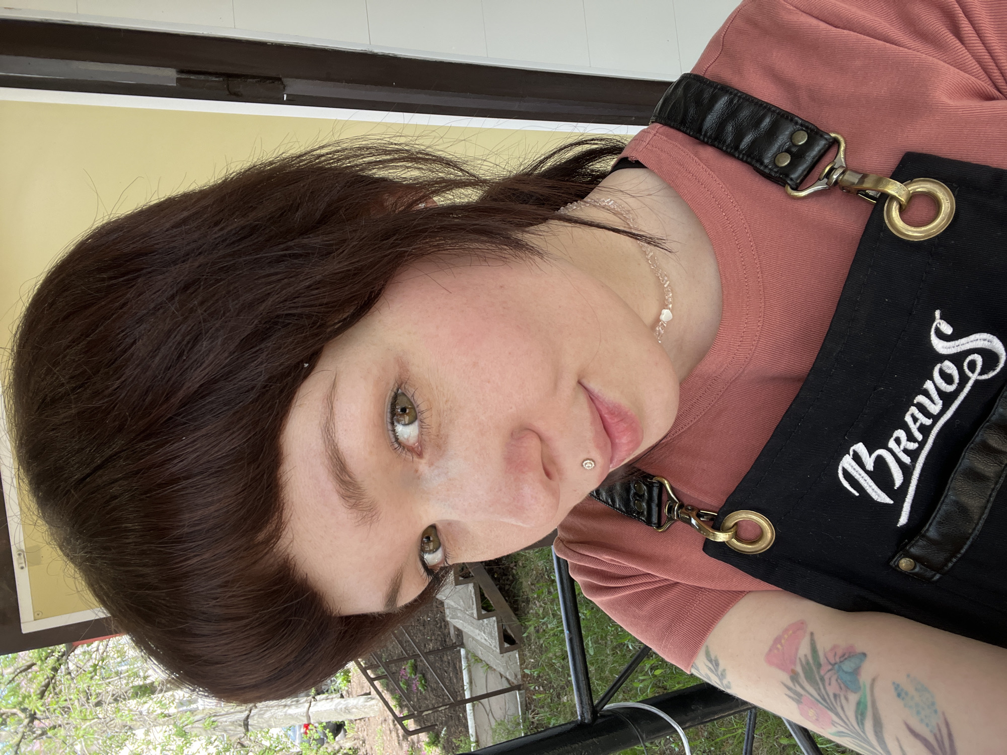 A medium close-up captures a woman wearing a pink t-shirt and a black apron with "Bravo" embroidered in white. Her dark hair is styled with a fringe. The woman has fair skin with freckles and her eyes are hazel. A small piercing is visible on the left side of her lower lip. She is also wearing a necklace. The woman is standing on an outside porch that has a black metal railing and a wood structure. The image appears to have been taken during the day, and the lighting is natural. A tattoo is seen on her left arm. The woman is outdoors in Chaikovsky, Russia.