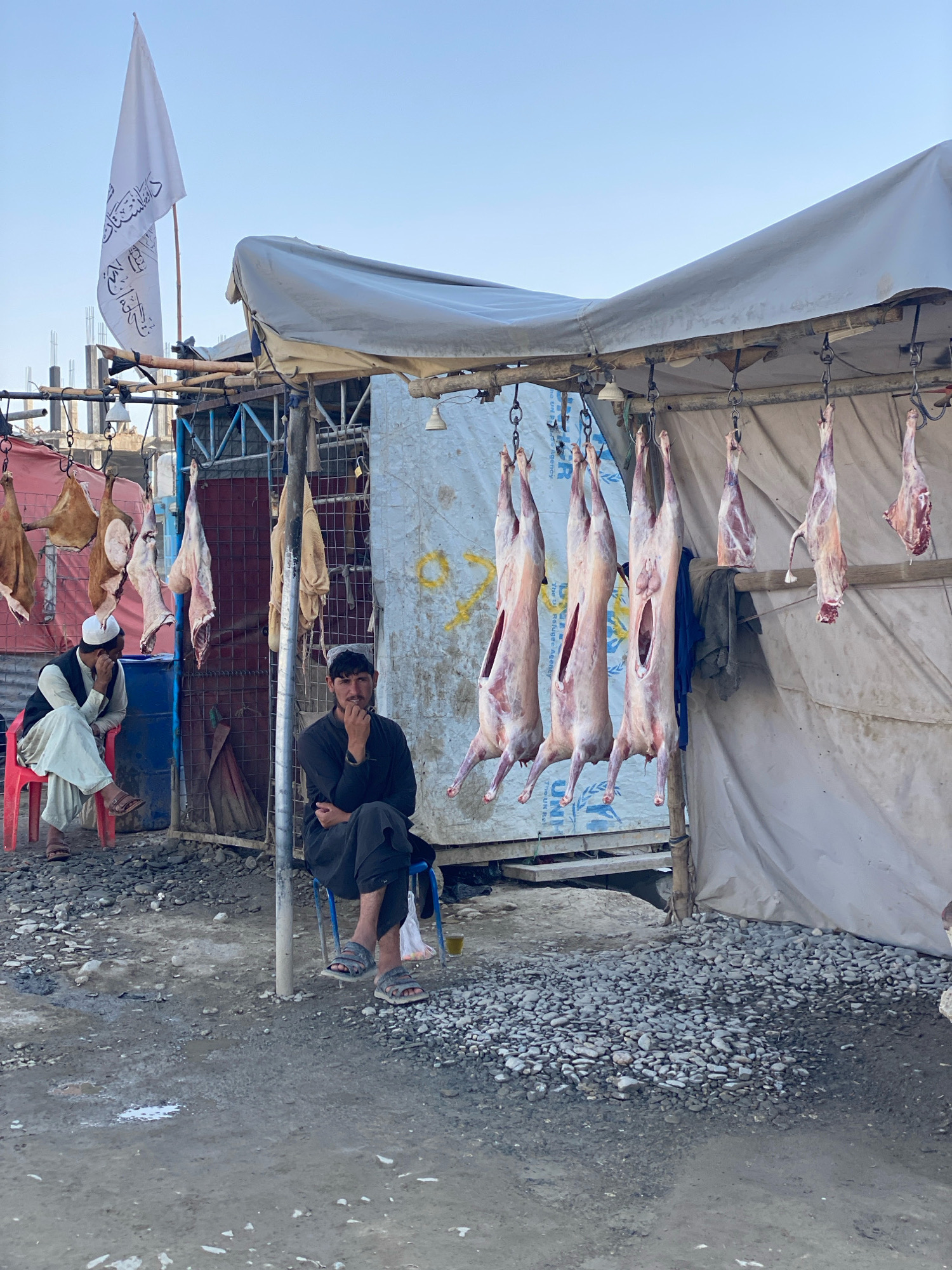 In Spin Boldak, Afghanistan, two men are seated at a roadside butcher stall. One man, wearing a white cap and traditional clothing, sits on a red plastic chair. The other man, in dark clothing, sits on a blue plastic chair. Both men appear to be observing the surroundings. The stall features several carcasses of sheep or goats hanging from a metal frame. The area is covered with gravel and dirt, and a white tent provides shelter for the stall. A white flag with Arabic script is visible in the background, along with some construction materials. The scene depicts a typical marketplace in the region.