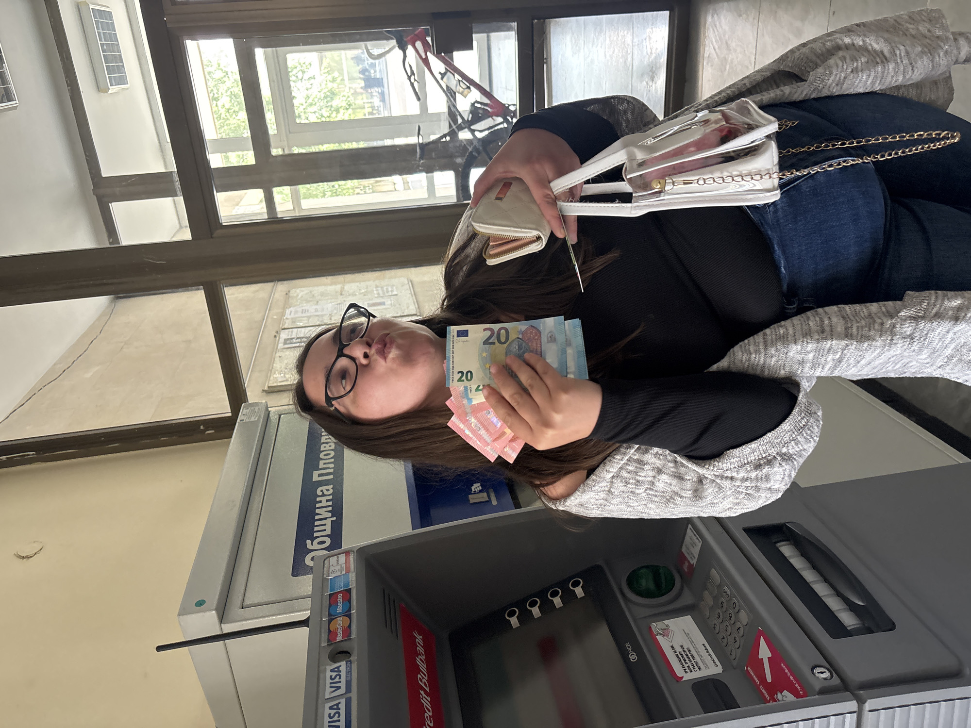 The image shows a woman standing next to an ATM machine in Plovdiv, Bulgaria. She is holding a stack of Euro bills in her hand. The ATM is branded with logos for VISA, MasterCard, and UniCredit Bulbank. There is text on the ATM in Bulgarian, including "община Пловдив" (Municipality of Plovdiv). The woman is wearing glasses and dark clothing with a gray sweater, and has a white wallet and handbag. There is a building with windows in the background, and a bicycle is visible through one of the windows.