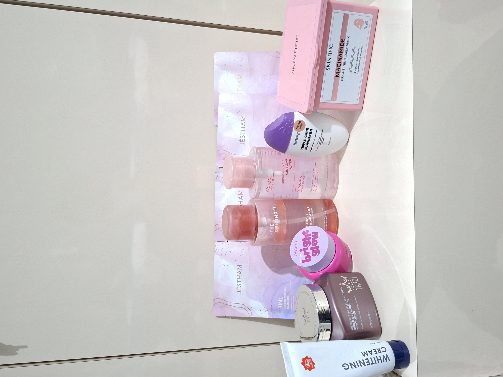 This photograph, likely taken in Kutaglumpang, Indonesia, depicts an assortment of skincare products arranged on a white, possibly melamine, surface. The items include a box of Skintific Niacinamide Brightening Daily Masks, Facetology Triple Care Sunscreen, Wardah Perfect Bright Micellar Water, The Originote Micellar Water, Emina Bright Glow Moisturizer, Tazzi Absolute Infused Whitening Night Lotion, Jestham facial masks, and Viva Whitening Cream. The products are displayed without any clear arrangement, suggesting a collection of personal care items. The environment appears to be a simple, indoor space, possibly a bathroom or vanity area, due to the presence of skincare products.