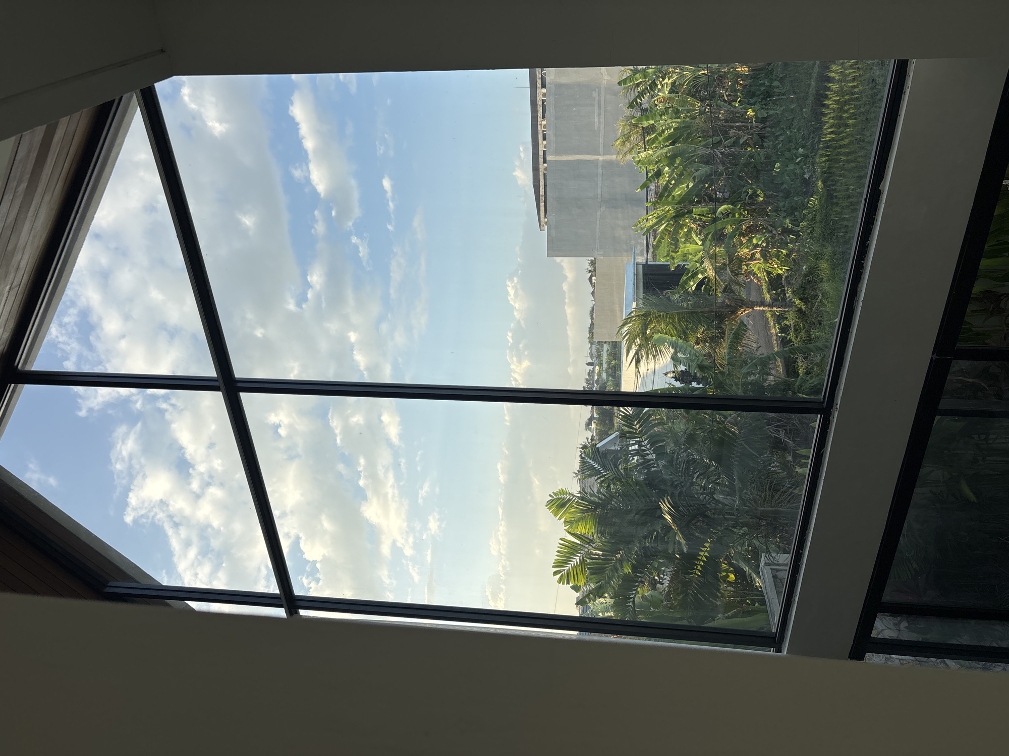 The image shows a view through a large, multi-paned window, likely from an elevated position within a building. The window frame is black. Through the glass, a tropical landscape is visible under a bright blue sky with scattered white clouds.
In the foreground of the outdoor scene, lush green vegetation, including palm trees and banana trees, is abundant. The sunlight casts long shadows, indicating it is either early morning or late afternoon.
In the middle ground, there are some buildings under construction. One prominent structure is a large, grey, unfinished building with visible concrete walls and a flat roof. Further in the distance, other residential or commercial structures can be seen, some appearing as white or lighter-colored buildings.
The overall impression is of a modern building overlooking a semi-developed area with abundant natural greenery in Mengwi, Indonesia. There are no people visible in the image.