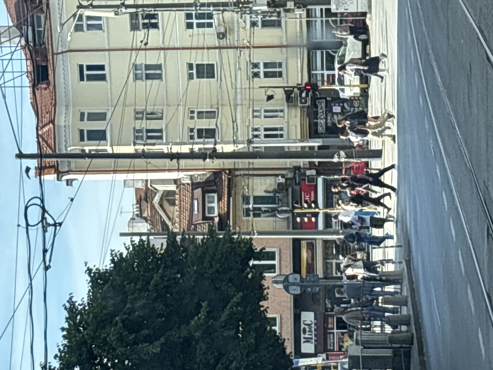The image depicts a street scene in Sofia, Bulgaria, on a bright day. In the foreground, there is a wide asphalt road with tram tracks. Several individuals are crossing the street at a designated crosswalk. On the left side of the road, a large, dark green tree stands prominently, partially obscuring a building with a shopfront displaying the "MADOC JEANS" logo. Adjacent to it is another building with a red awning. In the background, several multi-story buildings with terracotta tiled roofs and cream-colored facades line the street. One building has numerous windows with white frames. Electrical wires crisscross the sky above. Traffic lights are visible, indicating that the crosswalk is in active use. A white SUV is parked on the right side of the street, and more pedestrians are visible on the sidewalk. The overall atmosphere is that of a bustling urban environment during daylight hours.