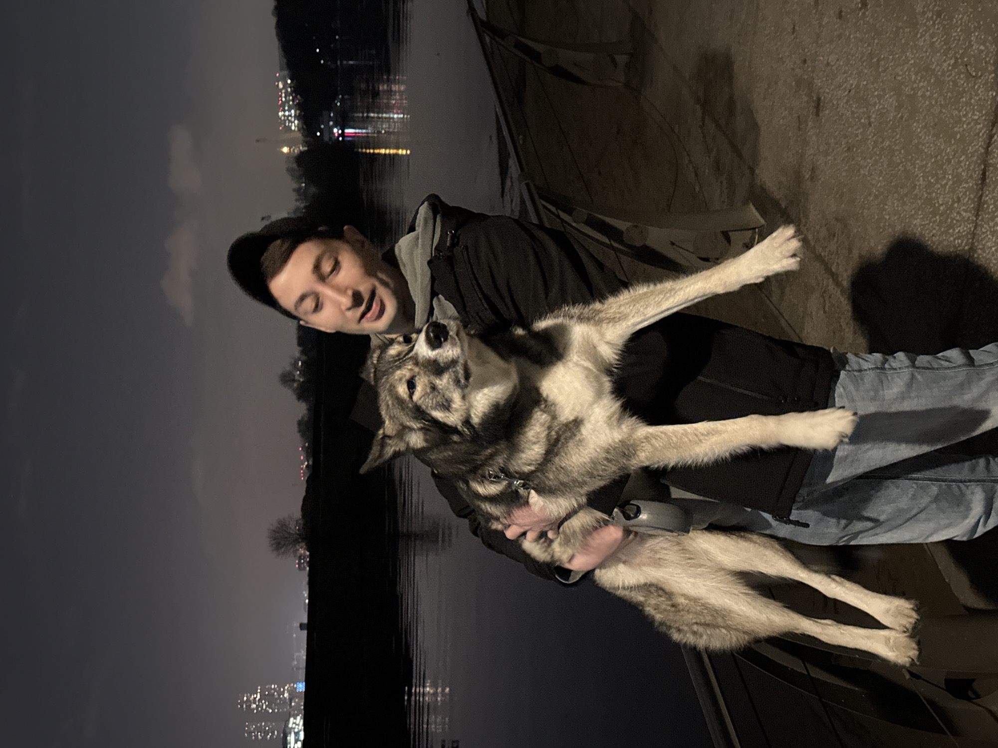 A man holds a large, wolf-like dog in an affectionate embrace at night by a body of water in Moscow, Russia. The man, with short dark hair and wearing a dark cap, a black hooded jacket, and blue jeans, smiles broadly with an open mouth as he looks at the dog. The dog, appearing to be a husky or similar breed with grey, black, and white fur, is suspended in the man's arms, looking up at him with an open mouth, appearing playful and engaged. A leash is attached to the dog's collar.
The scene is set outdoors at night, with the dark sky above. To the left, a river or canal reflects the distant, blurred lights of a city skyline. On the right, the man and dog stand on a paved embankment or pathway. Bare trees are faintly visible against the dark horizon across the water. The overall atmosphere conveys a warm, joyful interaction between the man and his pet. There is no visible text in the image.