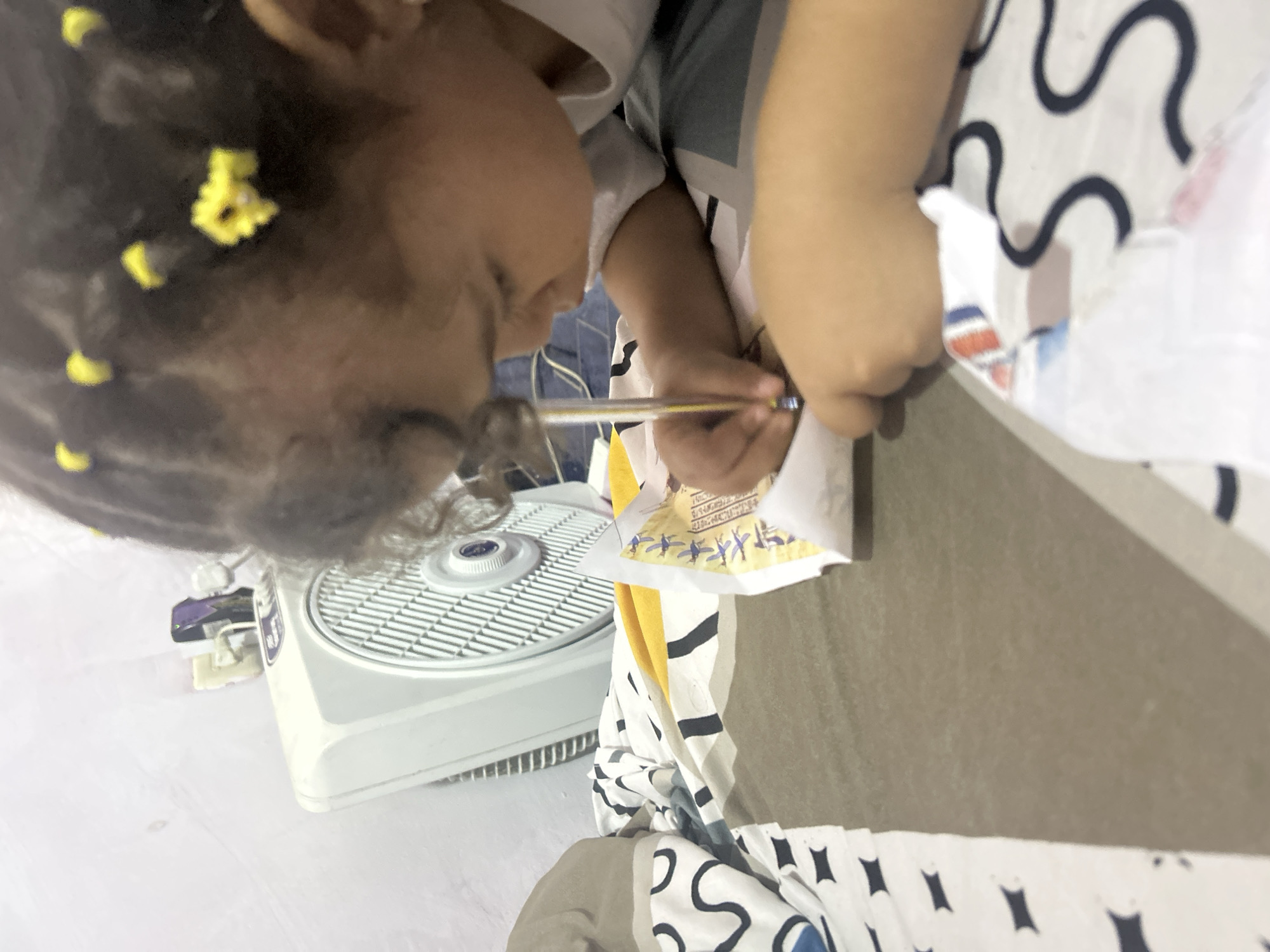 A young child with yellow hair accessories is holding a pen and appears to be writing on a piece of paper with images on it, positioned above a small white fan. The scene is indoors, and a patterned fabric is visible nearby. No specific location in San Juan de Los Morros, Venezuela can be determined from the image.