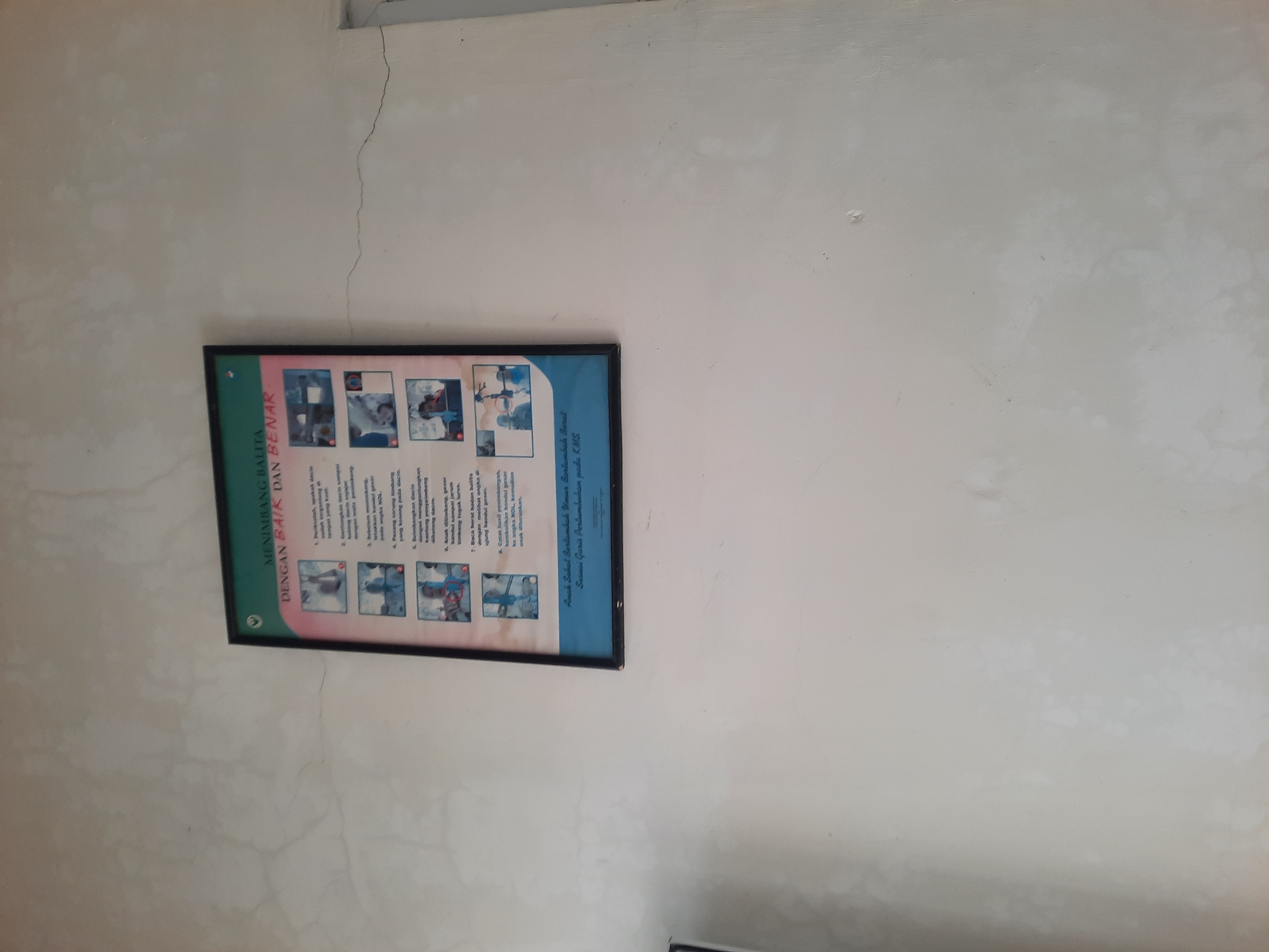 The image shows a poster mounted on a light-colored, textured wall. The poster, framed in black, has a green header with white text that reads "MENIMBANG BALITA DENGAN BAIK DAN BENAR". Below this, there are several small images depicting the correct procedure for weighing a toddler, along with accompanying Indonesian text describing each step. The poster is positioned slightly to the right of the center of the frame, and the wall appears to be an interior wall. The lighting suggests it's daytime. The overall environment seems to be a public space or health facility, likely in Lhokseumawe, Indonesia, given the Indonesian language on the poster.