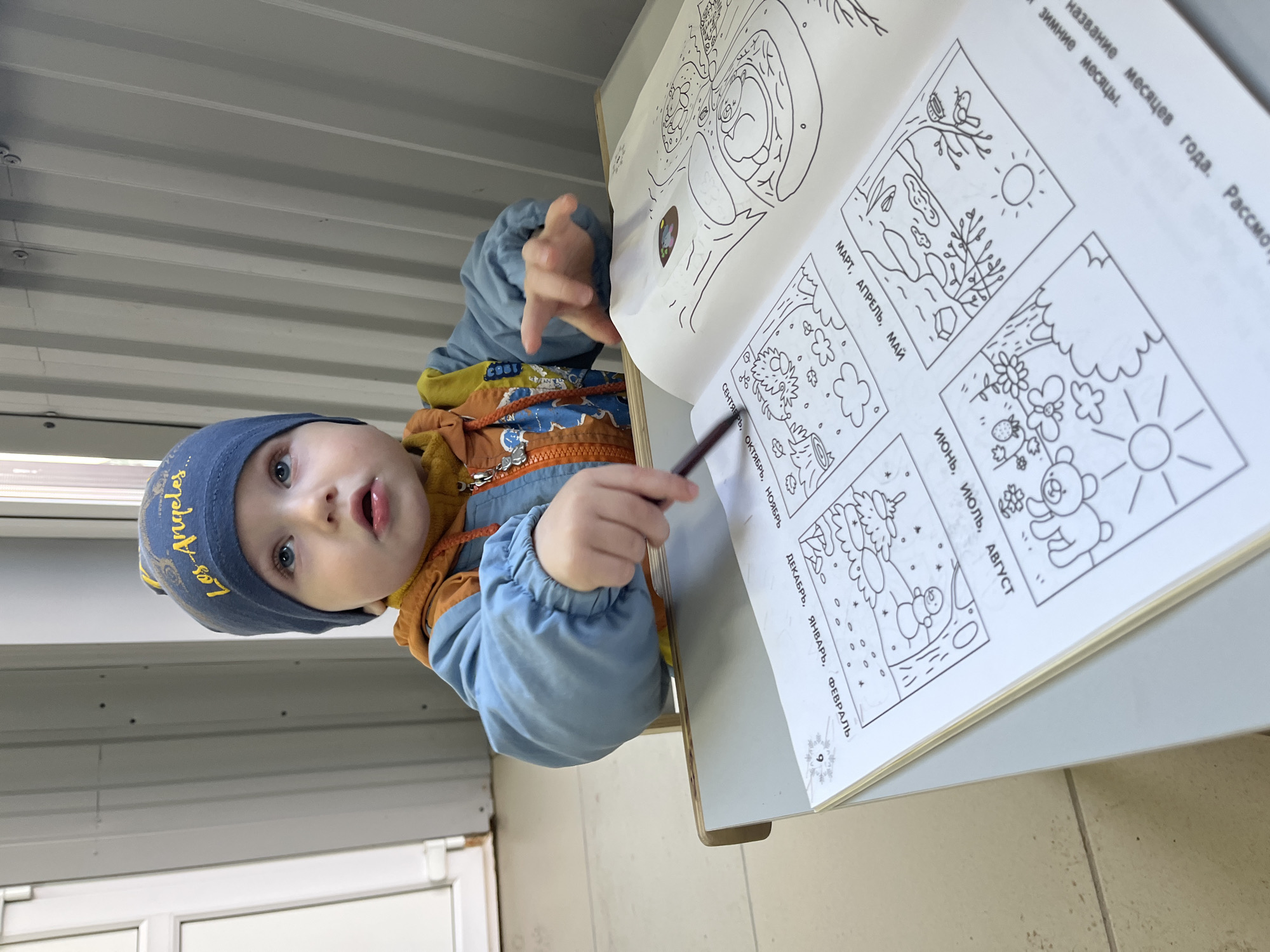 The image shows a young child, wearing a blue hat that reads "Los Angeles," a blue jacket, and an orange vest, standing next to a table. The child is holding a pencil and appears to be looking at and interacting with a coloring book open on the table. The coloring book features line drawings of different scenes representing the months of the year, with Russian text accompanying each image. The background suggests an indoor environment with corrugated metal walls and tiled floors. The photograph was taken in the city of Chaikovsky, Russia.