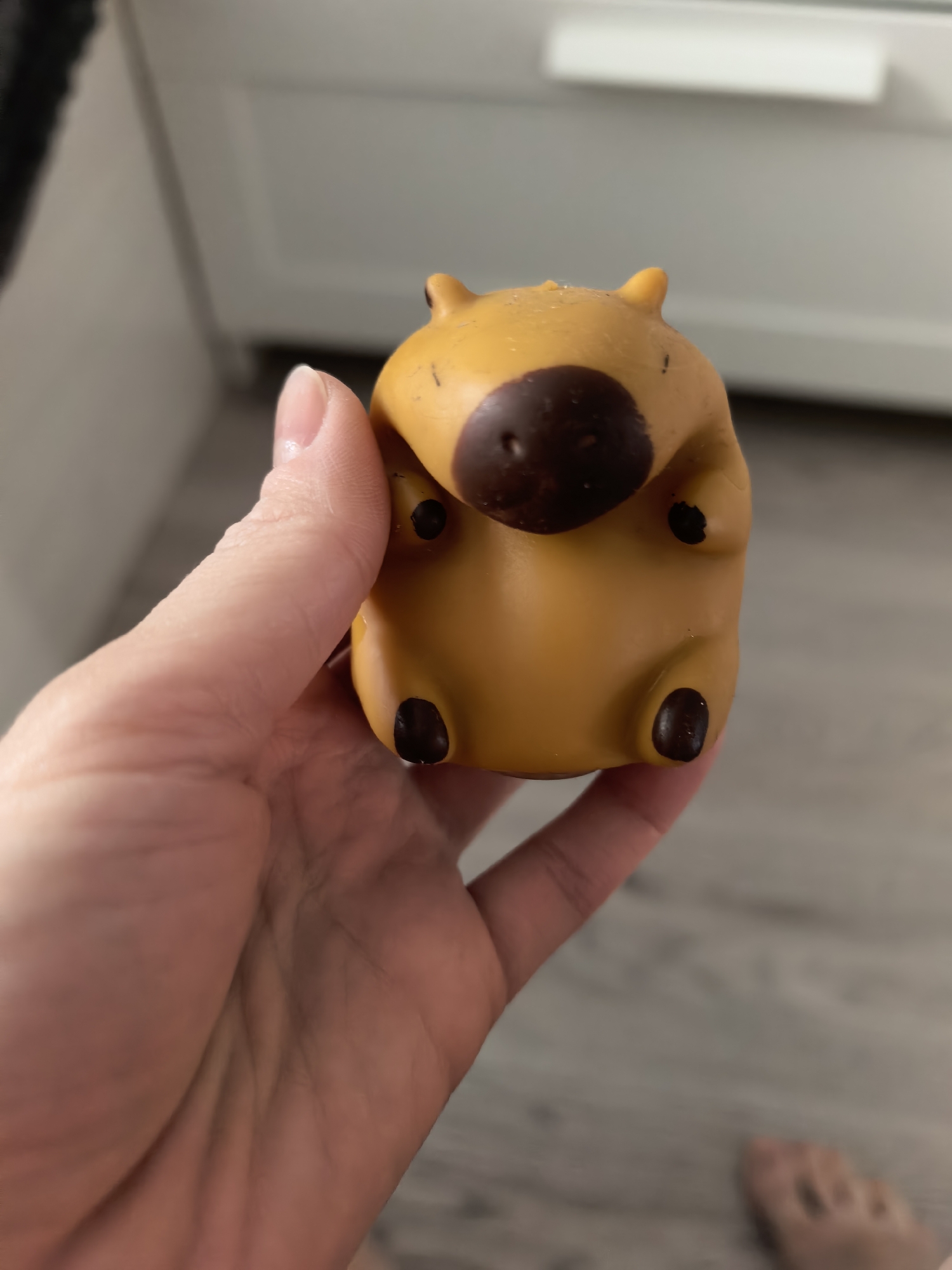 A hand holds a toy capybara. The toy is yellow with brown accents on the nose and feet. The background features a white cabinet and a gray wood floor. The setting is indoors and the location is unknown. Sterlitamak, Russia, is not evident in the image.