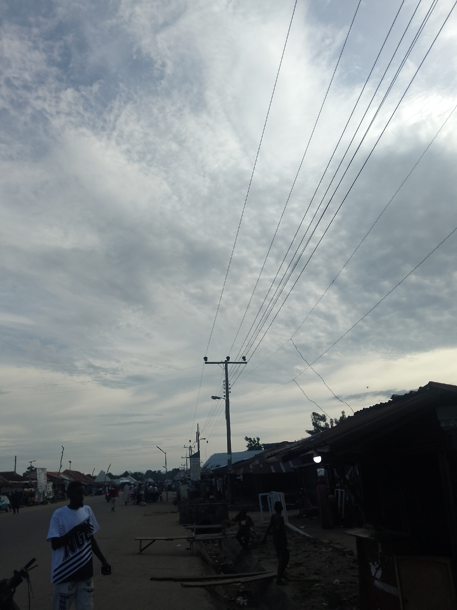 The image depicts a street scene in Serti, Nigeria, under a cloudy sky. In the foreground, a Black man wearing a white t-shirt with black and white diagonal stripes and patterned shorts stands on the left side of the road, holding a dark rectangular object, possibly a phone, in his right hand. He is facing slightly to his left, with his left arm bent and hand open. A portion of a motorcycle is visible in the lower left corner.
The street is lined with market stalls and buildings, some of which appear to be constructed with corrugated metal roofs. People are visible walking and gathered around the stalls. A utility pole with multiple wires extending upwards dominates the mid-ground. The road surface is unpaved and appears to be dirt or gravel. The overall impression is that of a busy, informal marketplace.
