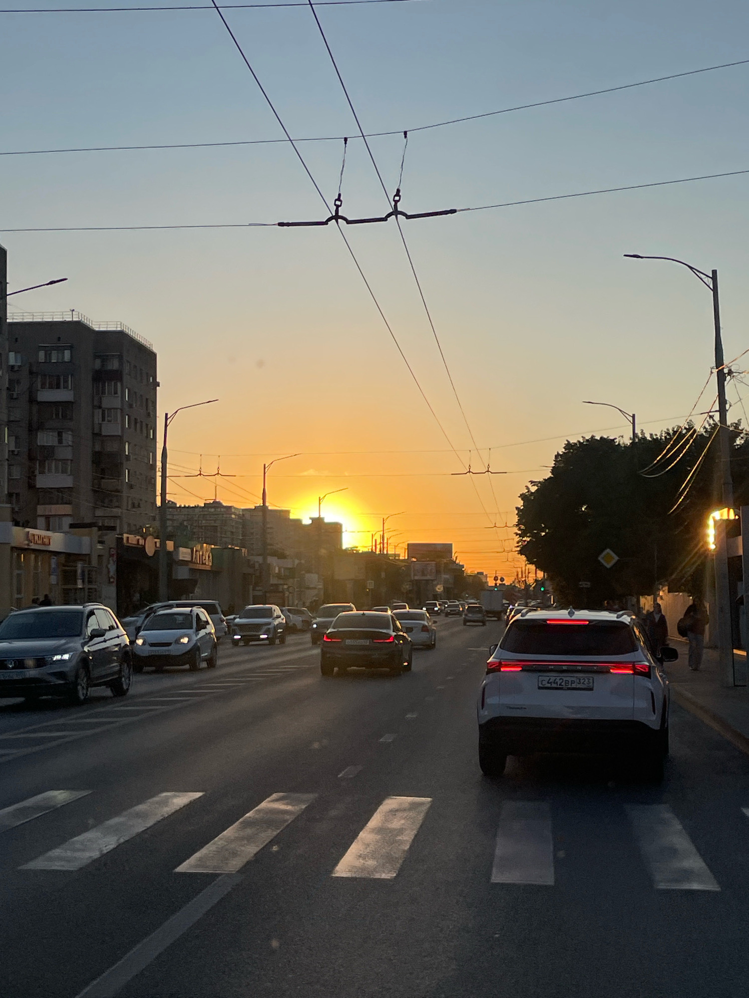 The image shows a street scene at sunset in Krasnodar, Russia. The sun is setting behind a line of buildings, casting a warm orange glow across the sky and the road. There are several cars driving on the road, which has pedestrian crossings marked with white stripes. Overhead, there are trolleybus wires and streetlights. Tall apartment buildings line one side of the street, and trees and smaller buildings are visible on the other. A few pedestrians are walking on the sidewalk. The overall impression is of a busy urban environment at the end of the day.
