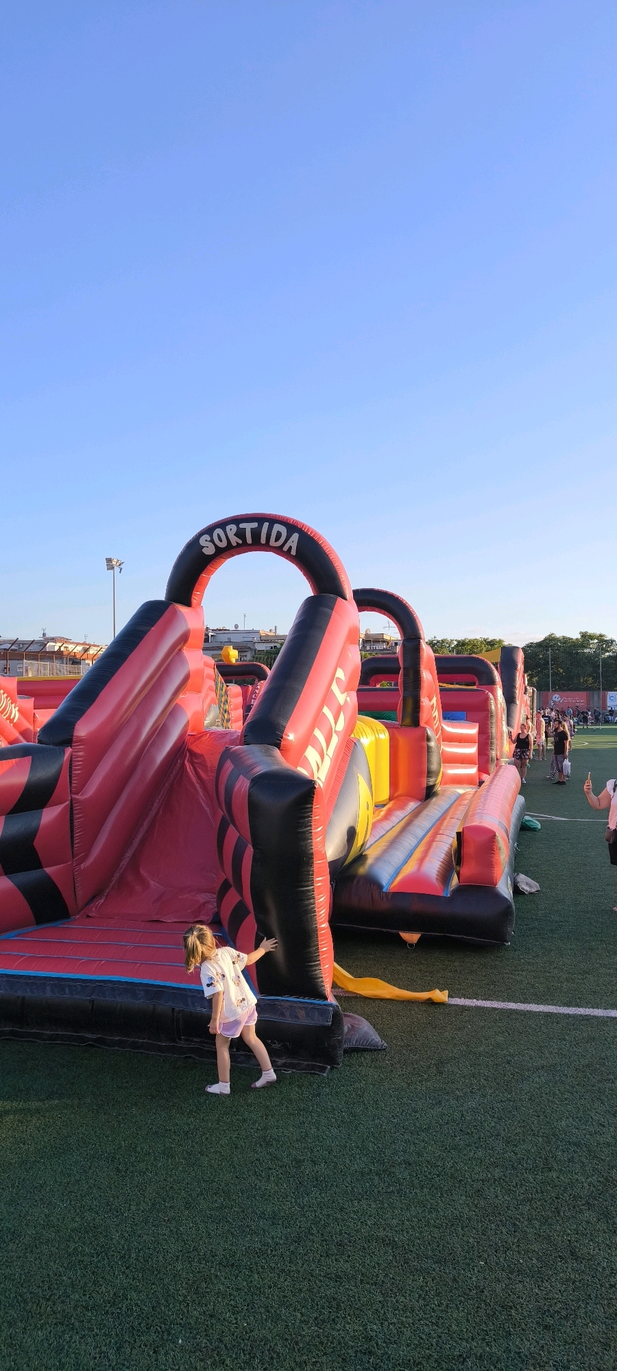 In Ripollet, Spain, an inflatable obstacle course with red and black sections is set up on a green turf field under a clear blue sky. The course features slides, climbing areas, and arches, one of which is marked with the word "SORTIDA" (exit in Catalan). A young girl in a white t-shirt with Mickey Mouse imagery and pink shorts is positioned near the base of the inflatable structure, touching it with her right hand and looking away from the camera. In the background, other people are visible, some walking and others standing, and structures that appear to be sports facilities are in the distance. The scene suggests an outdoor event or recreational activity.
