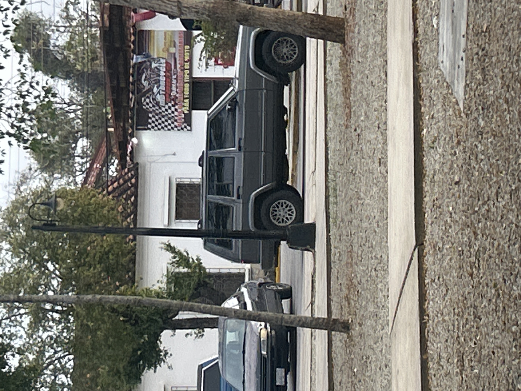 A gray Jeep Cherokee is parked on the right side of the street. The vehicle is facing right. In the background, there is a white building with a red tile roof. A banner with checkered flags and text is hanging on the building. A dark blue car is parked on the left side of the street, facing right. A narrow tree is in the foreground, to the left of the Jeep. The street is paved, and there is gravel on the side. The scene appears to be in Santa Ana, Venezuela.
