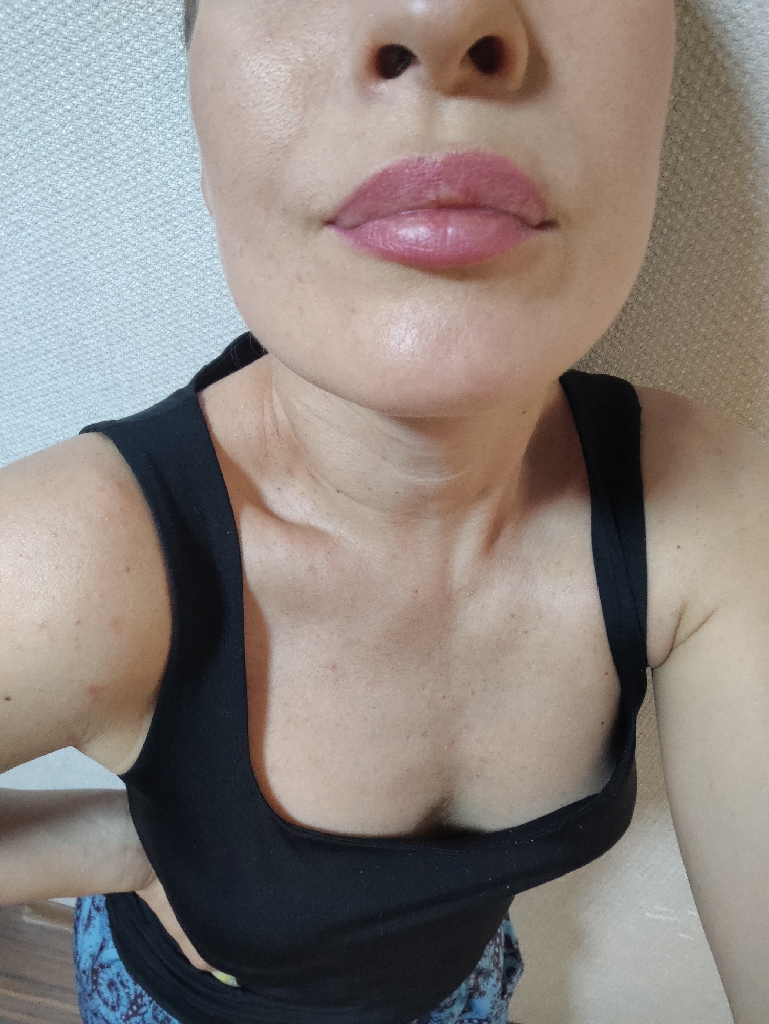 A medium shot shows a woman from the chest up, taken from a slightly high angle. She is wearing a black tank top and has on pink lipstick. Her skin has freckles and appears smooth. The background is a light-colored textured wall. The image does not contain any information about the city of Almaty, Kazakhstan, or any forensic details.