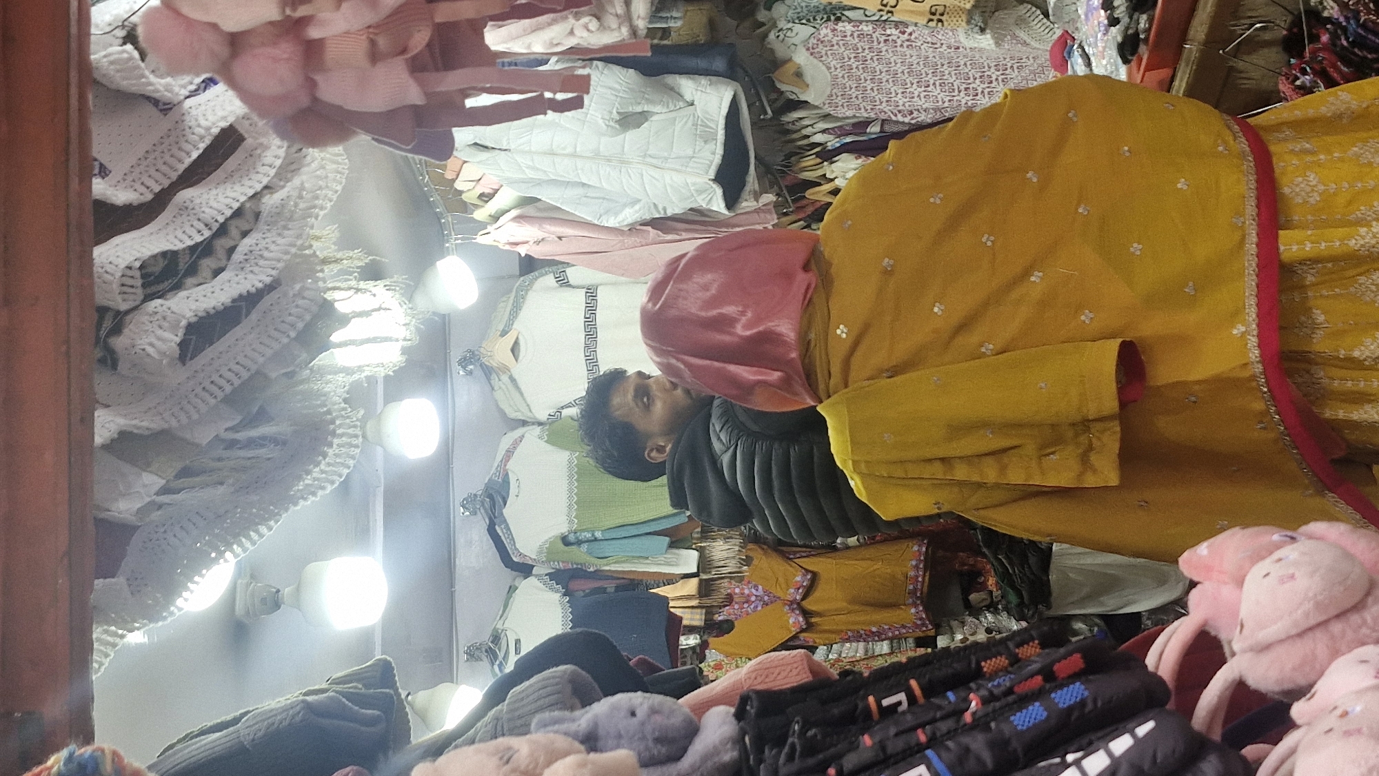 The image depicts the interior of a clothing store in Vashisht, India. In the foreground, a person wearing a yellow outfit with a pink head covering is facing away from the camera. To their left, a man in a black puffer jacket is looking to the side. The store is densely stocked with various garments, including sweaters, jackets, and other clothing items displayed on hangers. Overhead, decorative white knitted items with tassels are visible, along with ceiling lights. The scene captures a snapshot of a commercial setting with people browsing or working in a market environment.
