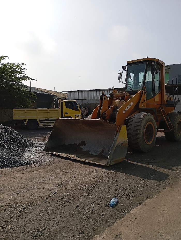 The image presents an outdoor industrial or construction material storage yard situated in the city of Situwangi, Indonesia, under a bright, overcast sky. The primary subject is a large, yellow Daewoo wheel loader positioned centrally, its front bucket lowered and resting near the unpaved, gravel-covered ground. Signs of dirt and use are evident on the loader's bucket and body. To the left and in the background, a yellow dump truck is parked. A dark, irregular pile of aggregates or gravel is visible on the ground to the left of the loader's bucket. The ground surface consists of compacted dirt and small stones, appearing dry. A single crumpled light-colored plastic bottle lies on the ground in the lower right foreground. The background consists of various industrial structures, including buildings with corrugated metal siding, and a tree with green foliage on the far left. No individuals are present in the scene, and no active work is depicted.