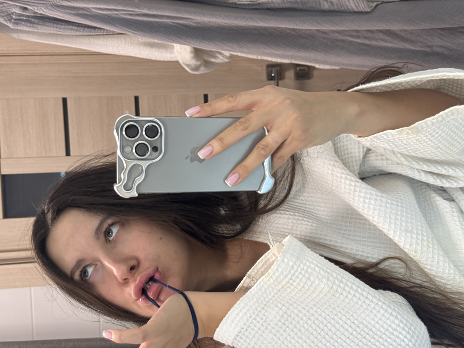 A young woman in a white waffle-weave robe takes a selfie with a grey smartphone. The phone has a silver, sculptural case and perfectly manicured nails are visible. She holds a dark blue hair tie in her mouth, eyes glancing upward. Behind her, a wooden door with dark vertical stripes and a metal towel rack are visible. The composition is off-kilter, with the background slightly blurred. Part of a rumpled, light grey blanket is present in the top portion of the image.