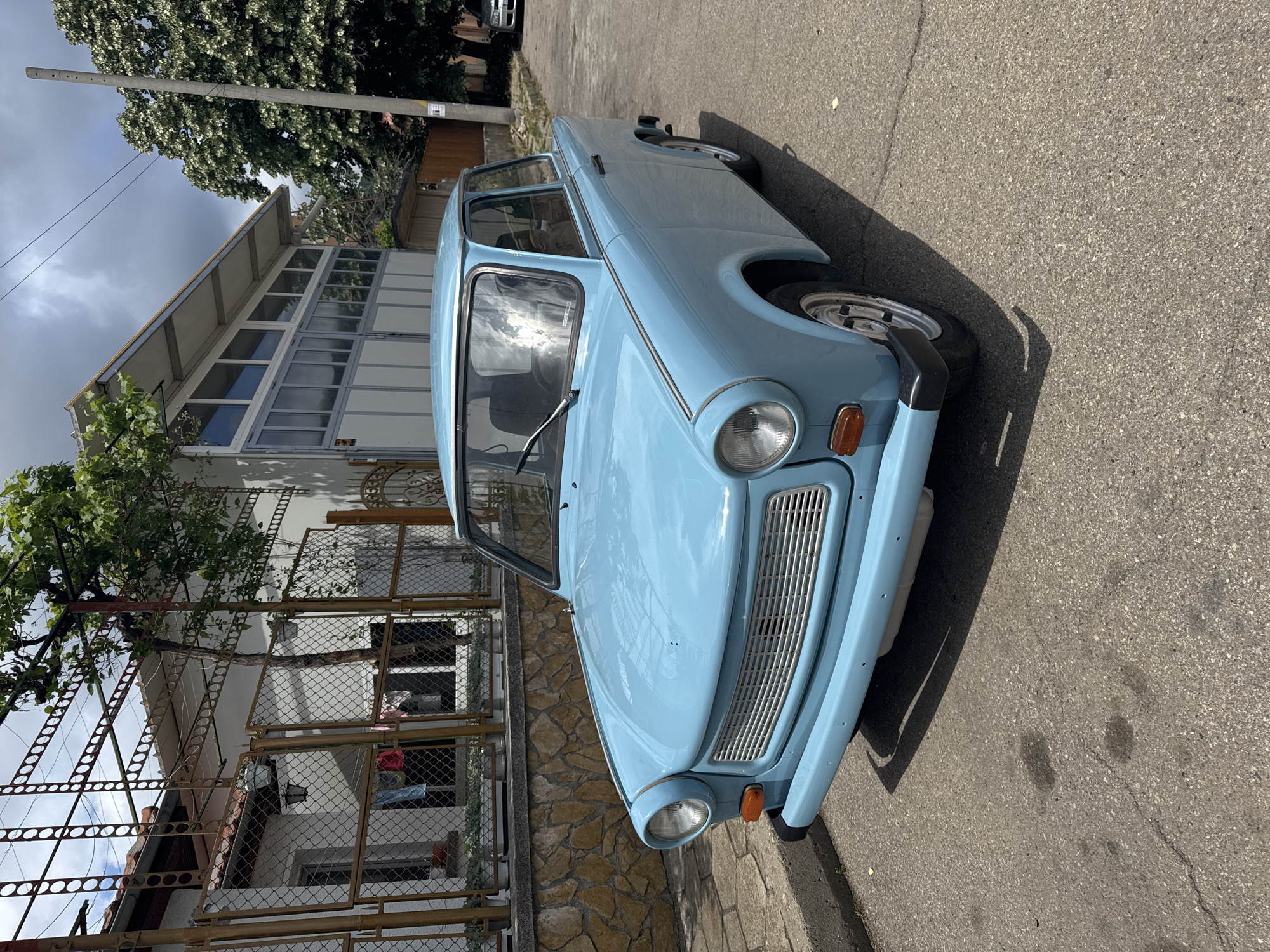 A light blue Trabant automobile is parked on an asphalt road in an unidentified city. The car is angled towards the camera, showcasing its front grill, headlights, and bumper. The background features a house with a distinctive A-frame architectural design and large windows. A fence and some vegetation are visible in front of the house. A utility pole stands to the left of the frame, adjacent to a tree with dense foliage. The sky is partially cloudy, and shadows cast by the car indicate a daytime setting.