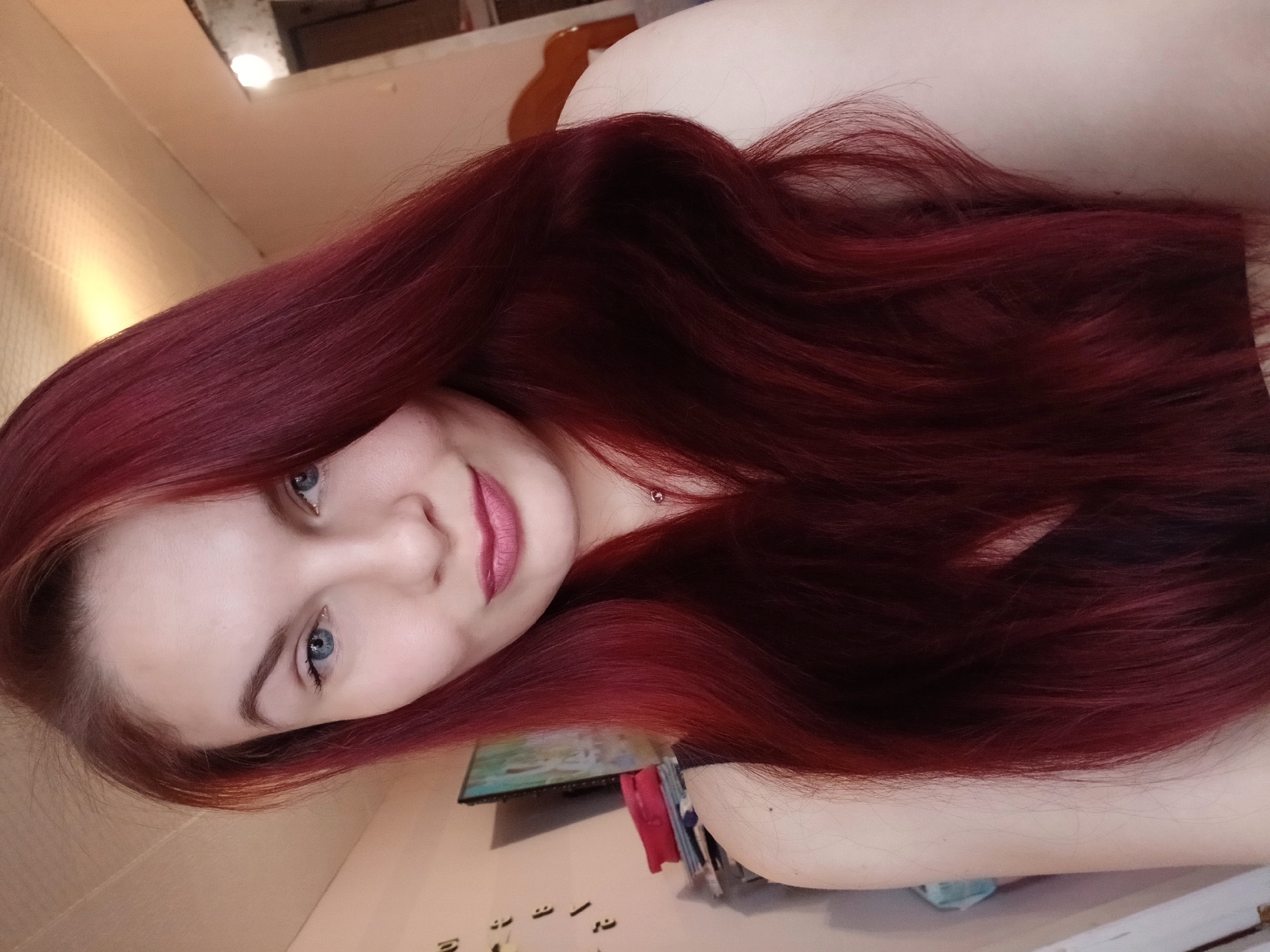 A woman with red-dyed long hair and blue eyes is angled towards the camera in what appears to be an indoor setting. The location appears to be in a room that is a bedroom or living space. The woman's face is visible, and the lighting in the room is bright.