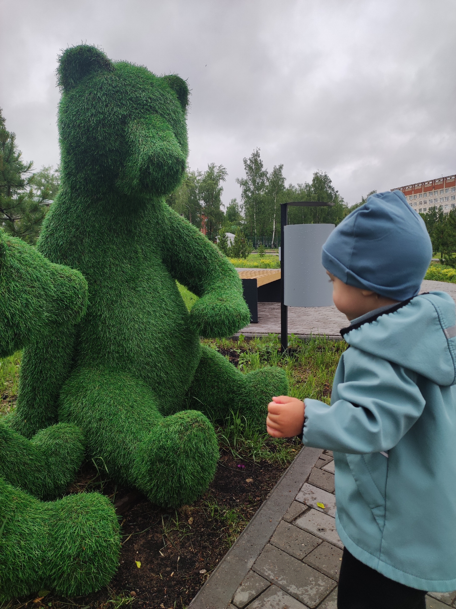 A young child wearing a blue jacket and hat looks at a large green topiary shaped like a seated bear. The scene is set in a park with trees and a building visible in the background. A paved walkway is in the foreground, and a trash receptacle and bench are visible behind the bear. The photograph was taken in the city of Chaikovsky, Russia. The weather appears overcast.