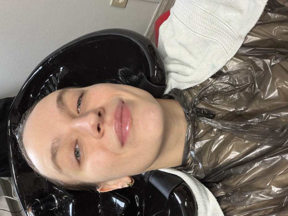 An adult female is depicted in a close-up, high-angle selfie within a hair salon, located in the city of Khimki, Russia. She is positioned reclining backward with her head resting in a black, glossy ceramic hair washing basin, typically used for shampooing or rinsing. Her hair appears wet and is slicked back into the basin. She wears a dark, translucent plastic cape secured around her neck, covering her upper torso and shoulders. A white, ribbed towel is draped over her shoulders, partially visible beneath the cape and over the edge of the basin. The woman has fair skin, blue eyes, light brown eyebrows, and is looking directly at the camera with a subtle smile. Her lips are slightly parted and appear to have gloss. A small gold hoop earring is visible on her left earlobe. The background consists of a plain light grey wall, with a light-colored electrical outlet or switch plate mounted on the right side. A small portion of a red chair is visible beneath the towel on the right side. The overall scene indicates an active hair treatment process.