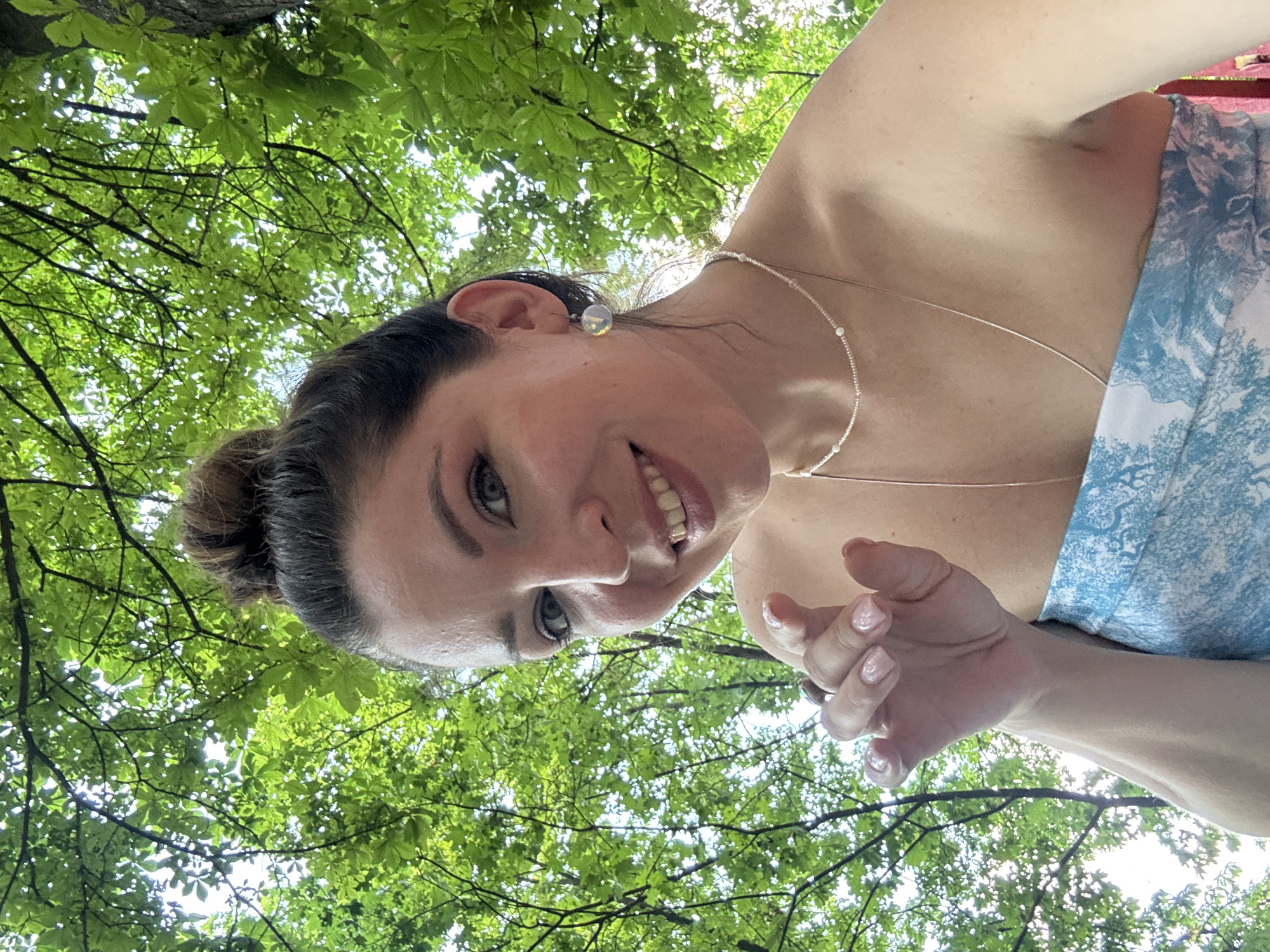 The media file shows a woman looking up at the camera from beneath a canopy of lush green leaves. She has her hair pulled up in a bun and is wearing a light-colored necklace and an earring. Her expression is a friendly smile, and her eyes are bright. The background is dominated by the vibrant green foliage of trees, with dappled sunlight filtering through the leaves, suggesting an outdoor setting on a clear day. The camera angle is from below, looking up towards the woman and the sky, creating a dynamic perspective. The woman is wearing a dress with a blue and white pattern. Her hand is visible in the foreground, with painted nails. The scene conveys a sense of natural beauty and joyful connection. The location context is Kryvyi Rih, Ukraine.