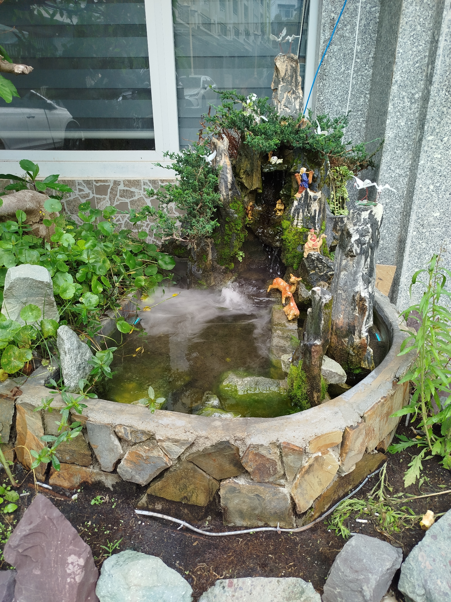 The image shows a small, ornamental fountain with a rock and cement base, set outdoors. Water appears to be circulating, creating a mist effect. The fountain incorporates figurines of people and animals, with small plants and moss growing on the rocks and wood features. The fountain is bordered by various plants and rocks. In the background is a building with a window, providing a partial reflection of a car. The image was taken in Da Lat, Vietnam.