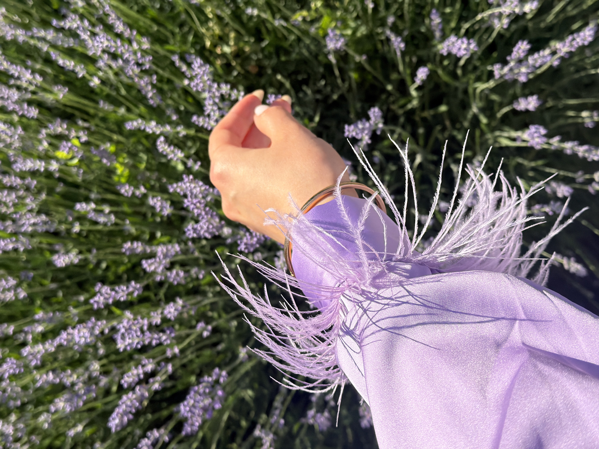 An overhead, slightly angled medium shot captures a person's left hand and forearm in the foreground, set against a blurred backdrop of lush, blooming lavender plants. The person wears a light purple, silky long-sleeved garment adorned with delicate, wispy white feathers around the wrist. A thin gold bangle is visible on the wrist. The hand is gently extended towards the lavender, with the index finger and thumb reaching out to lightly touch a flower. The background consists of dense clusters of purple lavender flowers interspersed with green foliage, bathed in bright, natural light that casts soft shadows. The overall scene evokes a sense of serene, outdoor exploration.