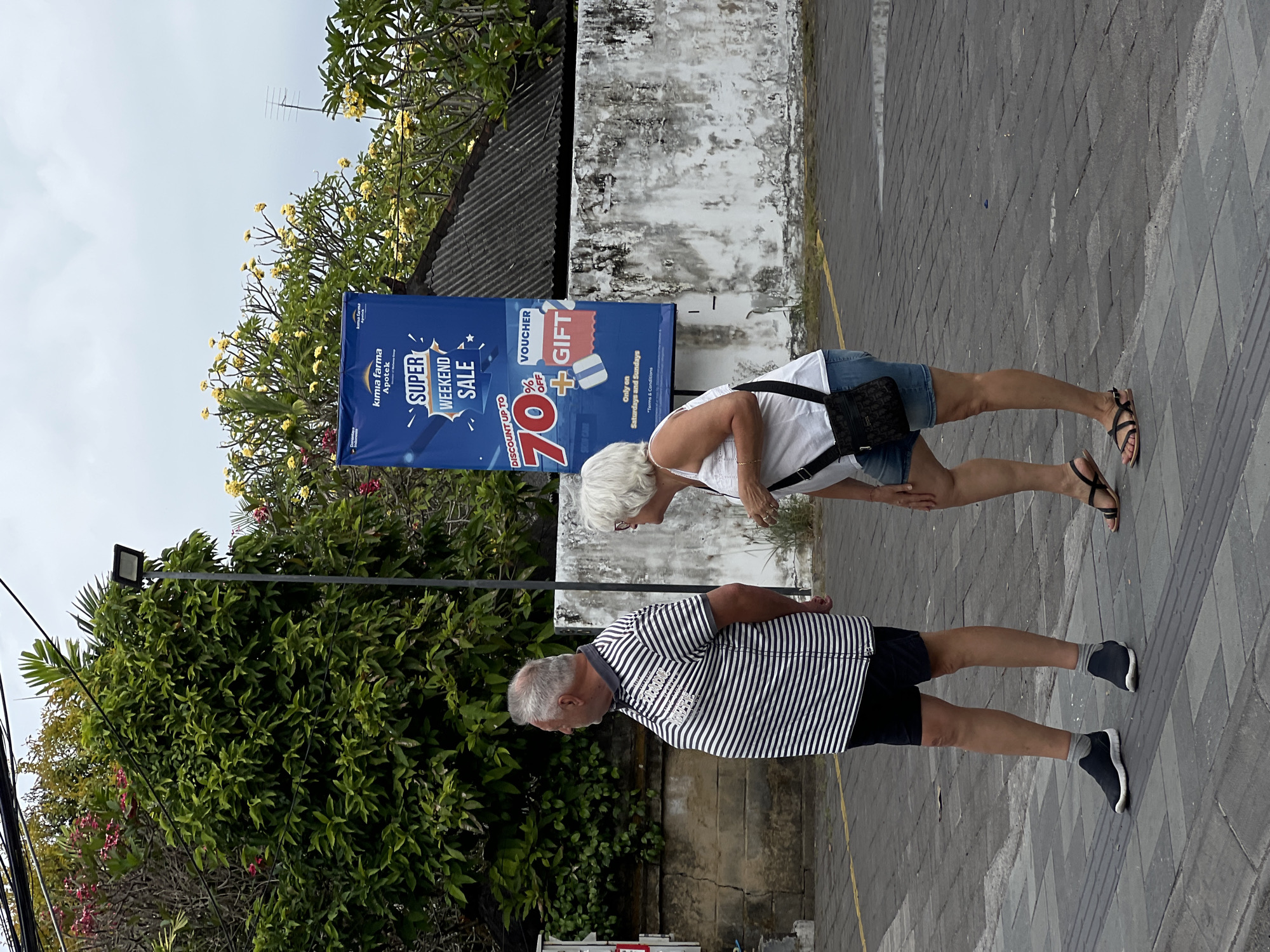 Two elderly individuals, a man and a woman, are walking on a paved sidewalk in Denpasar, Indonesia. The man is wearing a striped t-shirt and shorts, while the woman is dressed in a white top and denim shorts. They are walking away from the camera, with the man looking to the left and the woman looking forward. Behind them, a large blue banner advertises a "Super Weekend Sale" with a discount of up to 70% off and mentions vouchers and gifts. The banner is attached to a building with a white, weathered wall. Lush green trees with some pink and yellow flowers surround the scene, and overhead, power lines are visible against a cloudy sky. The environment appears to be an urban area, likely a street in Denpasar, given the presence of the banner for "Kimia Farma Apotek," a well-known Indonesian pharmacy chain. The ground is paved with grey tiles, and a yellow line is painted on the asphalt, possibly indicating a parking spot or lane.