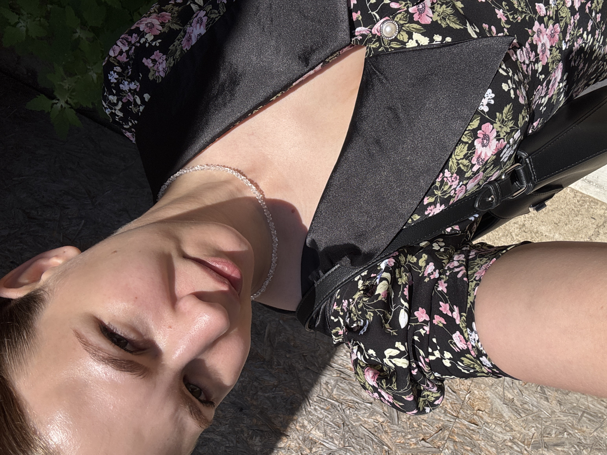 The image is a close-up, high-angle selfie of a young woman outdoors in Nizhnyaya Gar', Russia. She has fair skin and is looking towards the camera with a slight smile. Her hair is pulled back, and she is wearing a black floral print dress with puffy sleeves and a black collar. She is also wearing a delicate, clear beaded necklace. A black handbag with a strap is visible over her shoulder. The background consists of a rough, textured surface, possibly wood, with strong shadows cast by sunlight. Dark green foliage is also visible in the upper right corner.