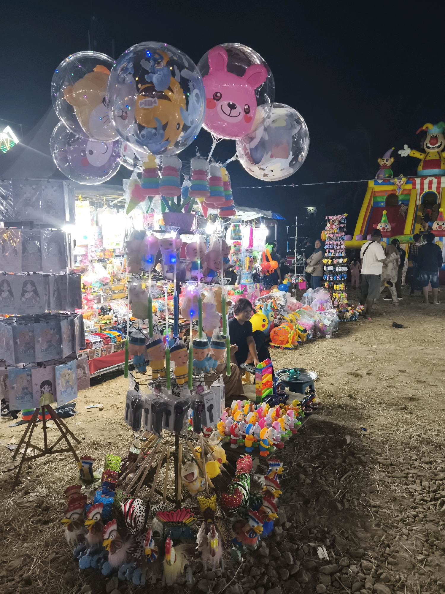 This image depicts a bustling night market scene, likely located in Palembang, Indonesia, characterized by its vibrant stalls selling a variety of toys and novelty items. In the foreground, a stall displays an assortment of colorful, rooster-shaped toys and other trinkets. Behind this, a larger stall features an array of animated toys with illuminated elements, and further back, a collection of large, transparent balloons with cartoon characters and LED lights are suspended.
Dominating the background is a large, inflatable playground structure, possibly a bouncy castle, adorned with colorful clown imagery. Various individuals are browsing the stalls and interacting within the market environment. The ground is a mixture of dirt and stones, indicating an outdoor, possibly temporary setup. The overall atmosphere is lively and festive, typical of local night markets.
