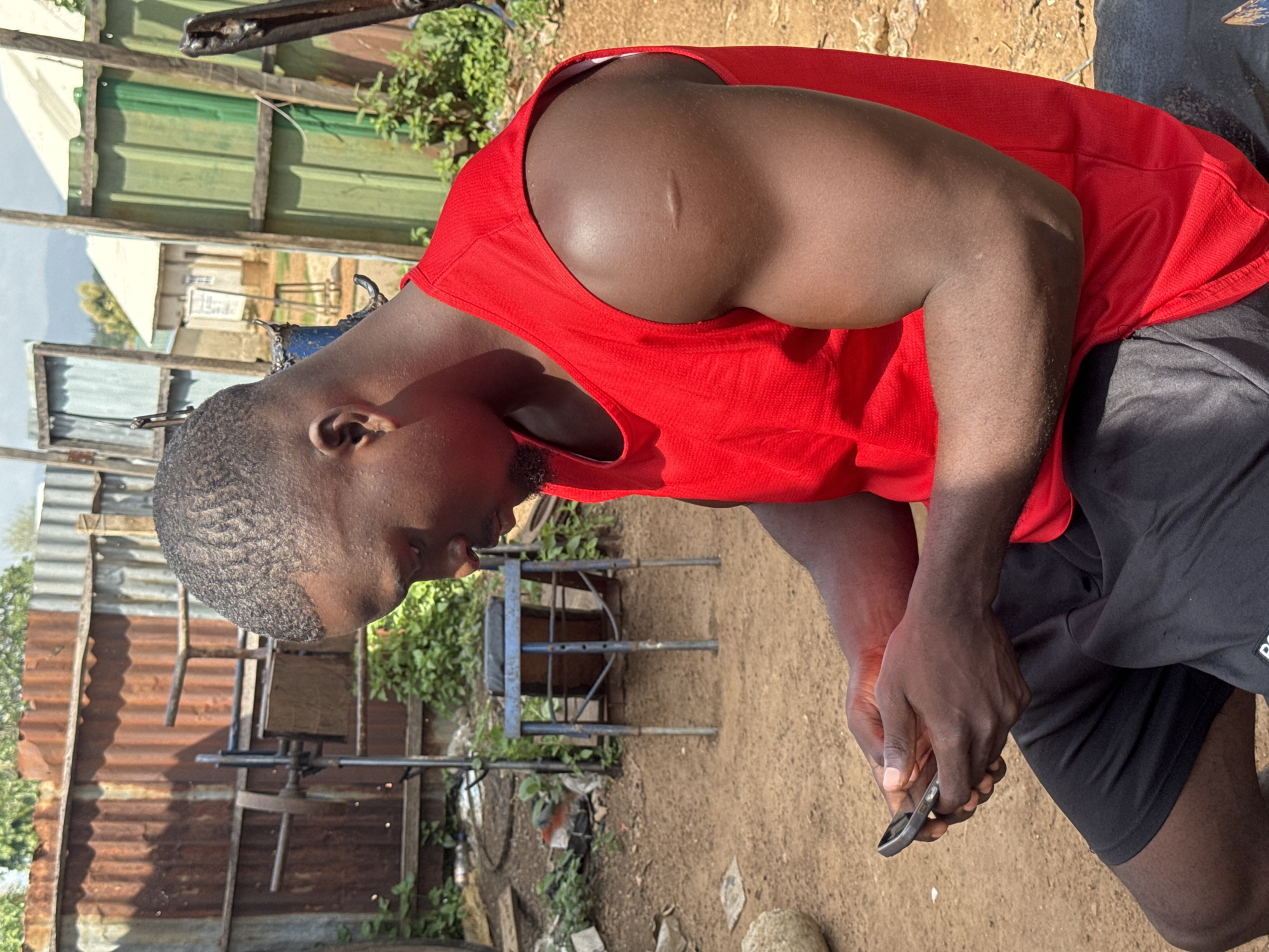 A young Black man with short, textured hair and a scar on his right shoulder is seated outdoors, looking down at a smartphone in his hands. He is wearing a sleeveless bright red shirt and black shorts. The environment appears to be an informal, possibly rural, setting in Akwanga, Nigeria, characterized by a sandy ground, overgrown vegetation, and corrugated metal fencing. In the background, there are structures made of corrugated metal and wood, and some distant buildings. The man is the primary subject and is engaged with his phone. The lighting suggests it is daytime.