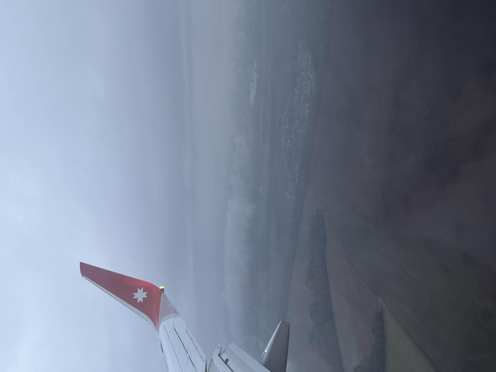 The image presents a view from an airplane window, showing the wing of the aircraft in the lower left and the ground below. The plane's wing has a red tip with a white star on it. The ground is partially obscured by a thick layer of fog or clouds, but fields and what appears to be a settlement are visible through the gaps. Based on the given context, the location is Новое Мартьяново, Russia.