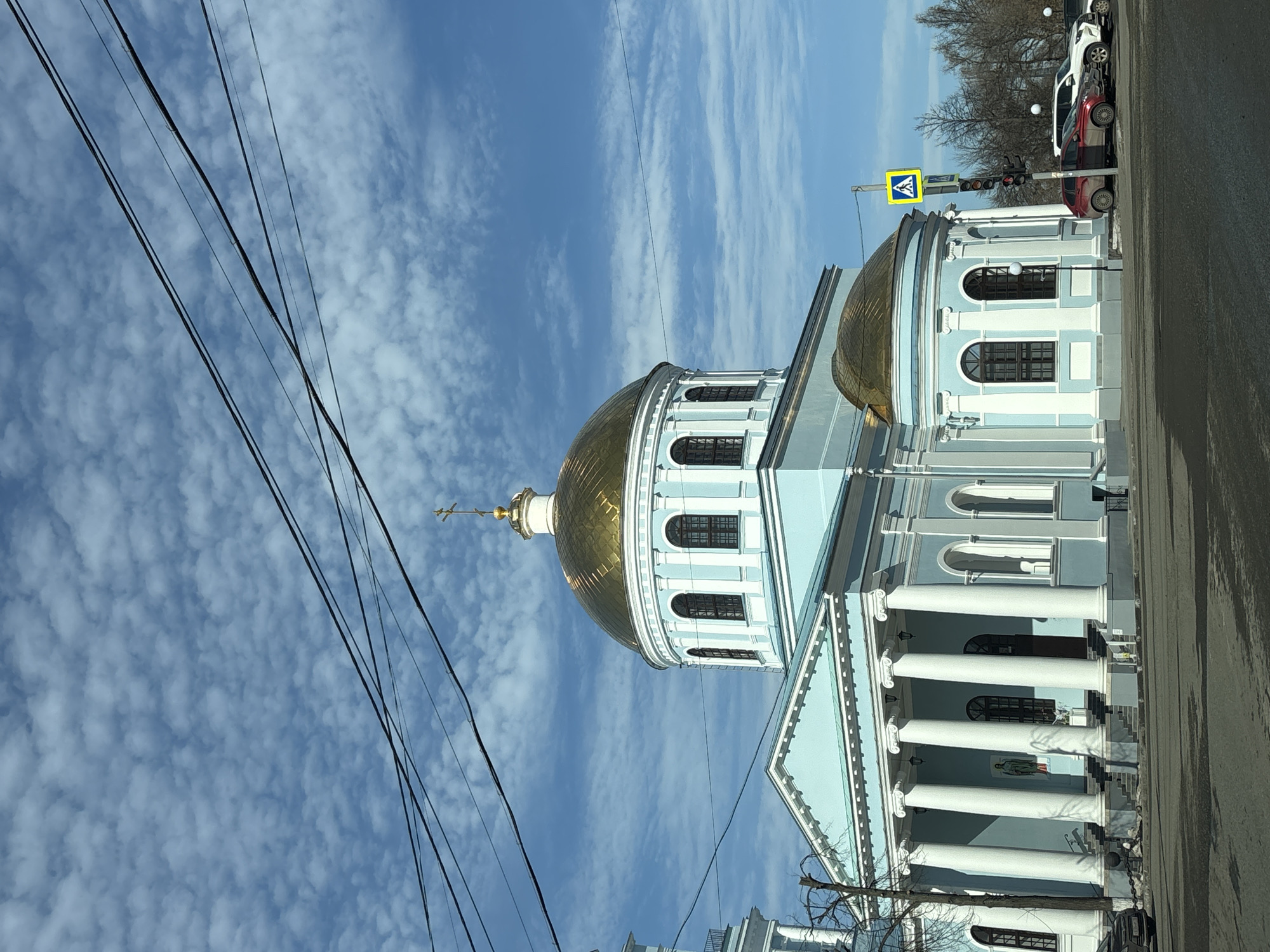 The image depicts the Ascension Cathedral (Voznesensky Sobor) located in Votkinsk, Russia. The cathedral is a large, light-blue Neoclassical structure with white architectural detailing, featuring a prominent central golden dome topped with an Orthodox cross. A smaller, golden dome is visible on a rounded side structure to the right. The main facade displays a portico with six tall, white classical columns. Numerous arched windows with dark frames are visible on the dome's drum and the building's body. A religious icon is affixed to the wall to the left of the main entrance, between two columns.
The foreground consists of a dark asphalt street, showing evidence of moisture or dirt. Overhead, multiple black utility lines crisscross against a bright blue sky populated by numerous small, white, scattered cumulus clouds. To the right of the cathedral, a yellow pedestrian crossing sign and a dark traffic light pole with a red traffic signal are visible. Several vehicles, including a red SUV and a white SUV, are parked parallel to the curb on the street to the right. Bare trees are visible in the distant background to the right. No individuals are discernible in the scene.