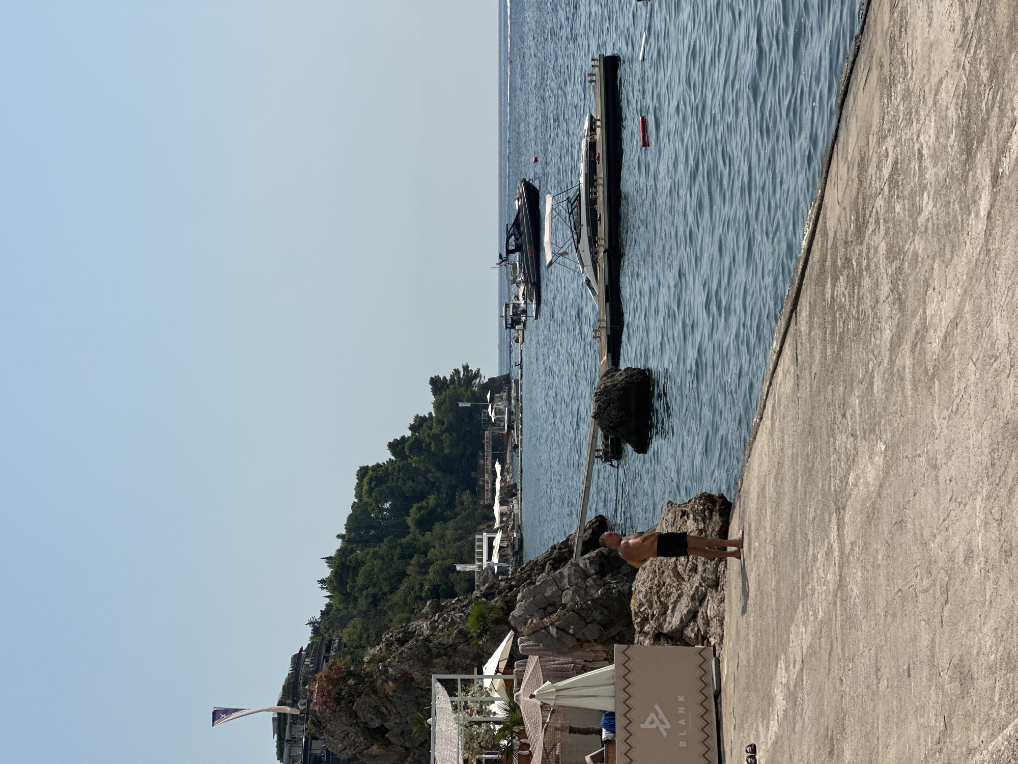 A man in black shorts stands on a concrete pier facing the Adriatic Sea. Behind him, a rocky cliff is covered in lush green foliage and dotted with buildings. Several white umbrellas and lounge chairs are visible on a beach area to his left, part of the Blank Seaside establishment. A wooden dock extends into the blue water, with a white speedboat moored to it. Further out, a larger dark-colored yacht is anchored. The sky is clear and blue, indicating a sunny day. The overall scene depicts a serene coastal resort setting in Budva, Montenegro.