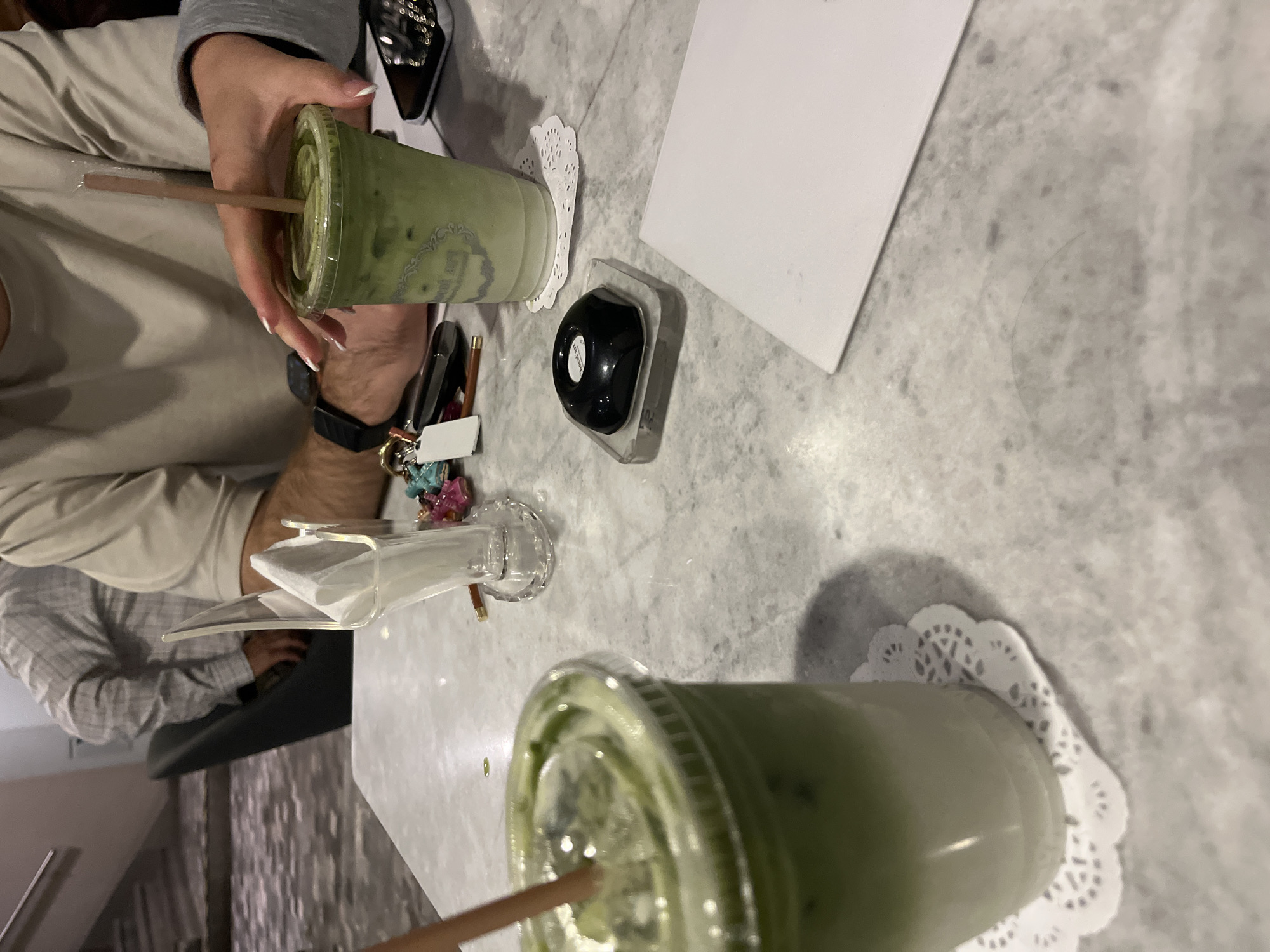 The image depicts an indoor scene at a Choc-Art cafe located in Puebla, Mexico, specifically identifiable by the "choc-art" branding visible on a drink cup and a small table object. The focal point is a light gray and white marble-patterned table.
Two individuals are partially visible at the table. On the right, a person's hand with manicured fingernails (French tip style) and a gray long-sleeved garment is reaching for an iced green matcha latte in a clear plastic cup with a lid and a light brown straw. This cup bears the "choc-art" branding and rests on a white paper doily. To the left of this person's hand, a forearm with visible hair and a black smartwatch is seen, belonging to another individual wearing a light beige short-sleeved shirt. This individual's hand is near a set of keys with multiple colorful keychains and a small white rectangular tag.
Two iced green matcha lattes are present on the table. One is in the foreground, left of center, also in a clear plastic cup with a lid and a light brown straw, resting on a white paper doily. Both drinks exhibit a layered appearance with a white base and a green top, indicative of a matcha latte.
Other objects on the table include a clear acrylic napkin holder with white napkins positioned centrally. To the right, a black smartphone is partially visible. Further right, a small black, glossy, dome-shaped object with "choc-art" written in white text rests on a clear rectangular base. A folded or stacked white paper object is also on the right side of the table.
In the blurred background, a third individual is partially visible from the back, wearing a light-colored, possibly checkered or striped long-sleeved shirt, seated in a dark chair. A staircase with a handrail is discernible in the far background on the left. The ambient lighting appears soft, consistent with an indoor establishment.