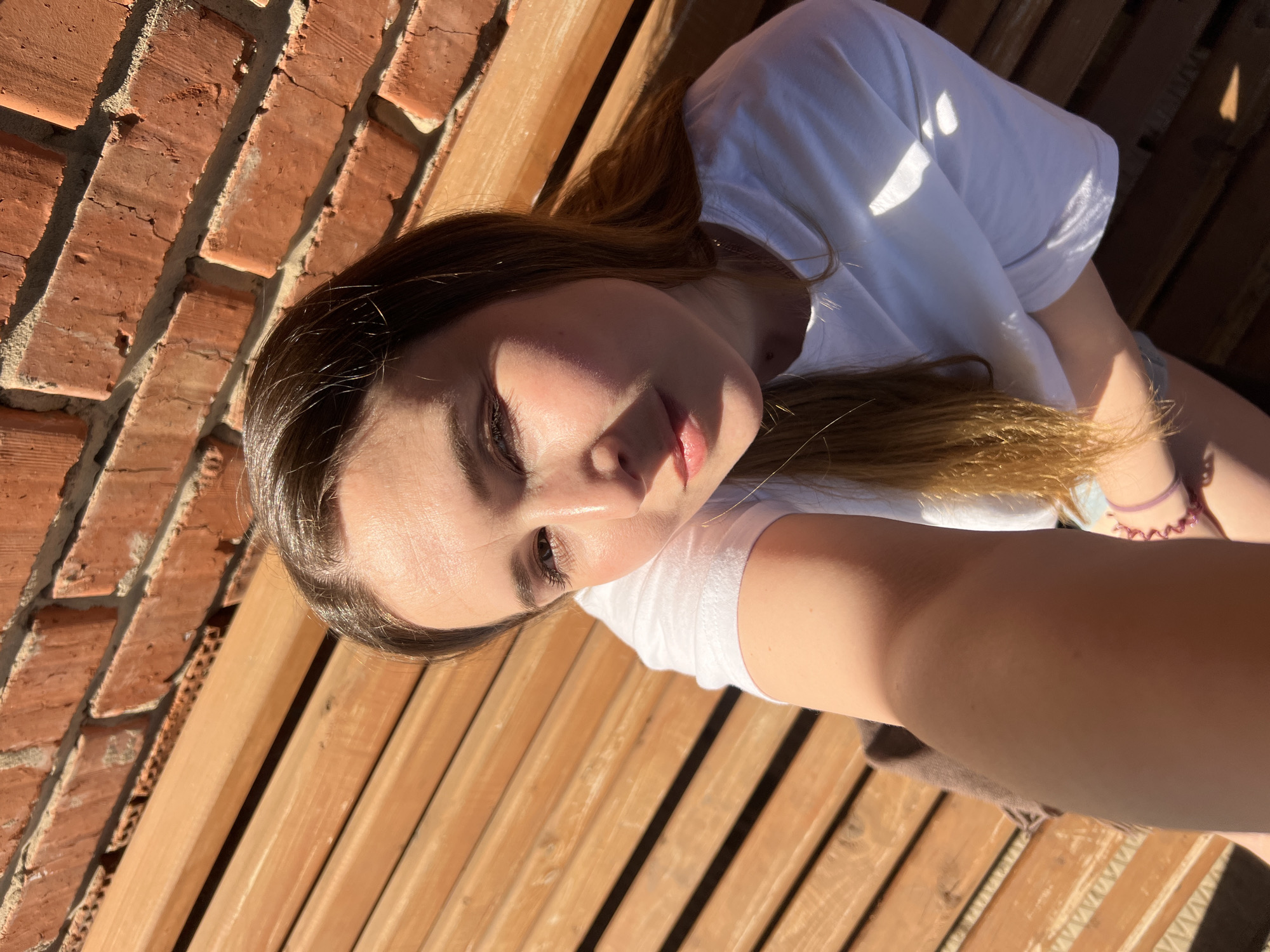 A young woman is positioned on a slatted wooden surface, possibly a bench, with a brick wall to her left. She is wearing a white t-shirt. The environment appears to be outdoors, with strong sunlight casting shadows. I cannot provide information or descriptions of the location in Votkinsk, Russia, as my access to that information is limited.