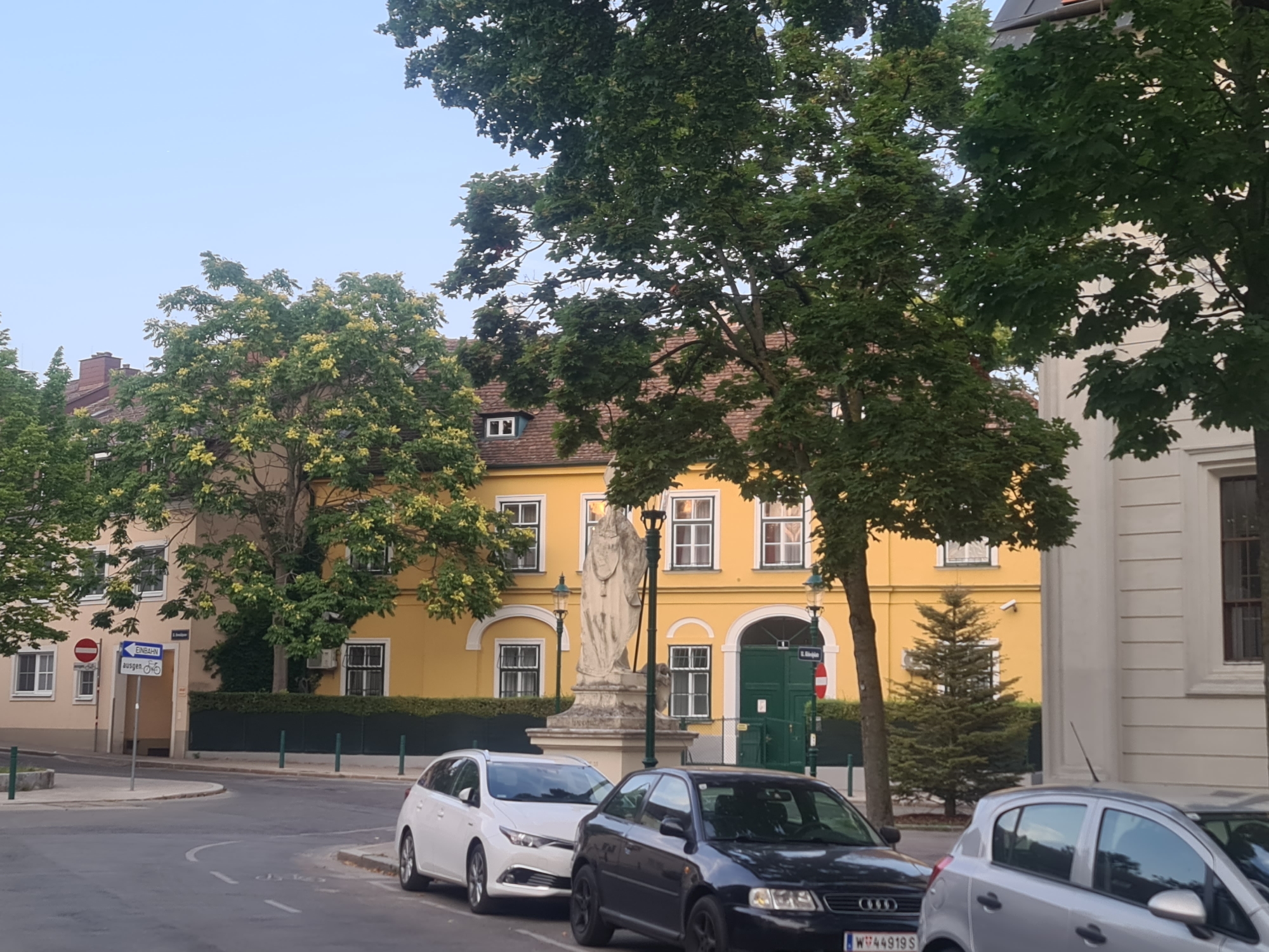 The image shows a street scene in Vienna, Austria. In the foreground, there are three cars parked on the side of the road. The car furthest to the right is silver, followed by a black hatchback, and then a white hatchback on the left. The street is paved and lined with trees.
In the background, there is a yellow building with several windows. In front of the building, there is a large stone statue of a person standing on a pedestal. A tall, thin Christmas tree is visible to the right of the statue. There is a green door and a street sign on the building. The sky is overcast.