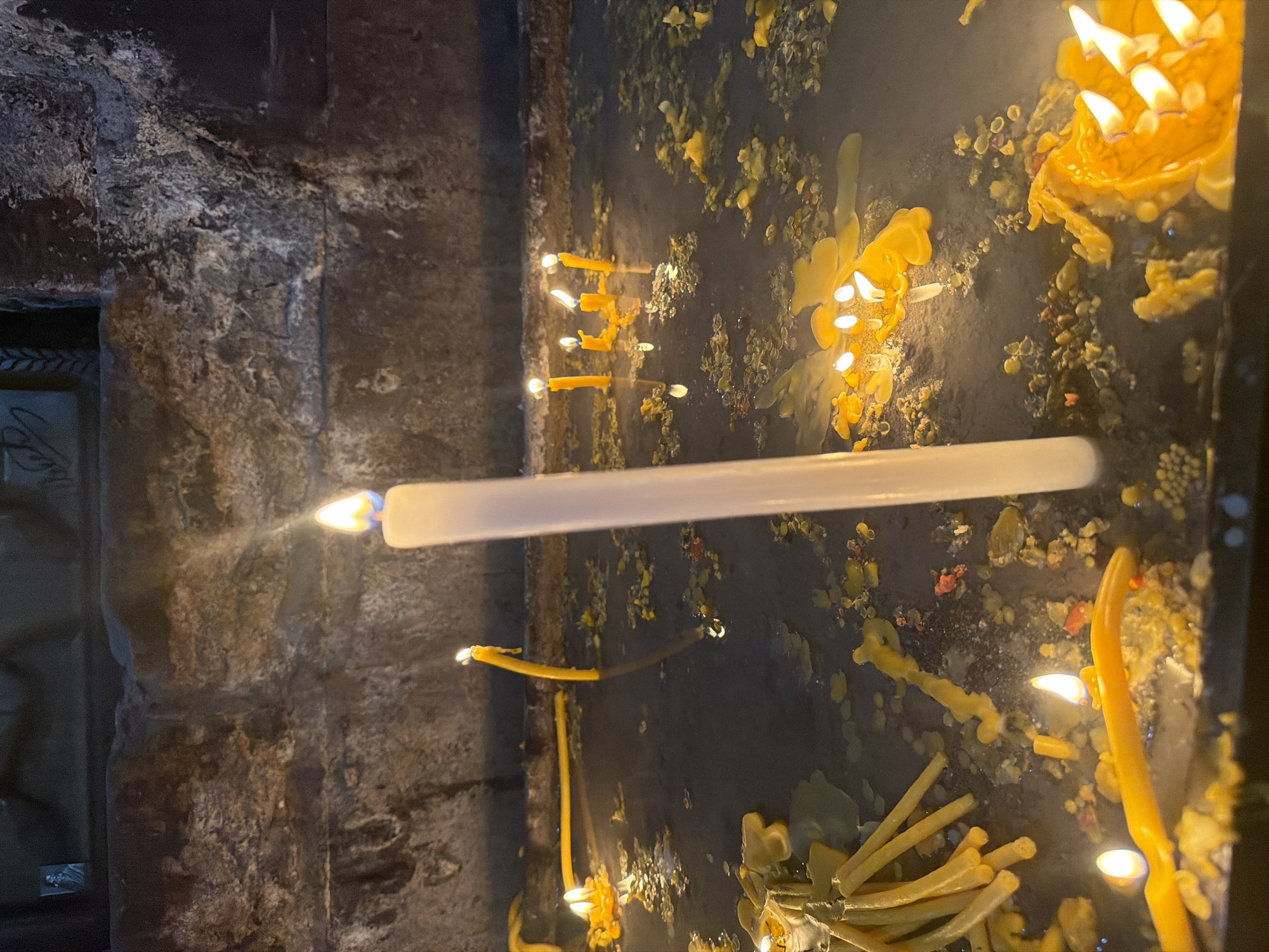 The image depicts a close-up view of a stone niche in a dimly lit environment, likely a place of worship. Several lit candles are present, with a prominent white candle standing upright in the center, casting a warm glow. The surface around the candles is covered in melted wax, forming pools and drips of various yellow and orange hues. Several unlit yellow candles are clustered on the left side of the frame, some appearing to be partially melted. In the background, a textured stone wall with visible imperfections and slight discoloration is present. The overall scene is suggestive of a devotional practice, possibly in the city of Ashtarak, Armenia, known for its historic churches and religious sites.