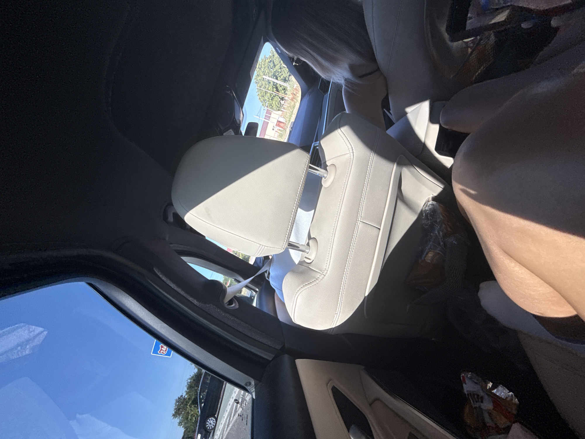 The image depicts the interior of a car, viewed from the passenger seat. The car is in motion, as evidenced by the road signs and trees visible through the windows, suggesting a highway or major road. A blue and red highway sign with the number "240" is visible on the left side, indicating the location is likely near a route numbered 240. The car's interior is clean and modern, with light-colored leather seats. A person's legs are visible in the foreground, indicating they are a passenger. A person with blonde hair is seated in the back seat, facing forward. The atmosphere is sunny, with clear blue skies. The overall scene suggests a road trip or travel by car.