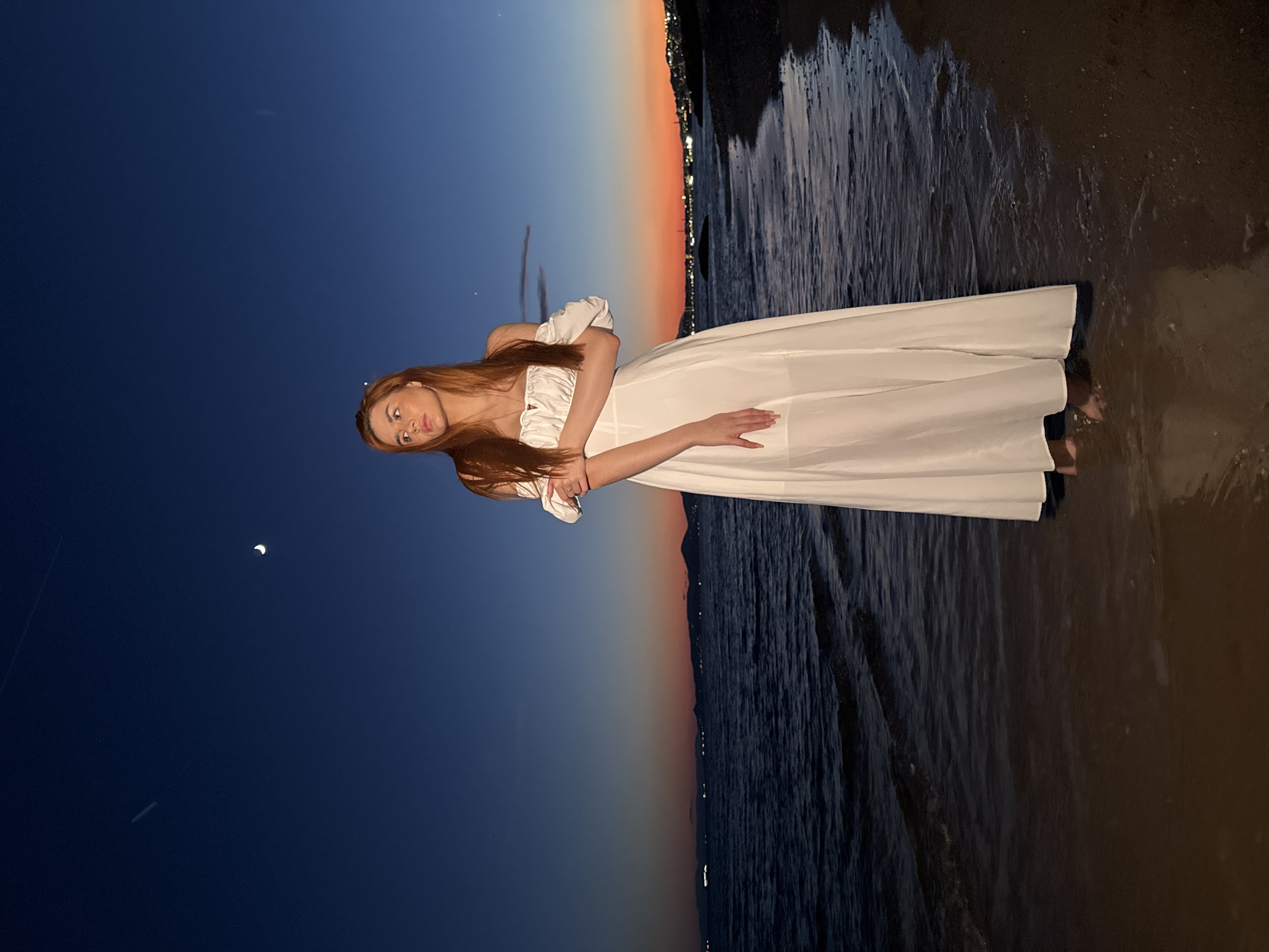 A young woman with auburn hair stands at the water's edge on a beach at dusk. She is wearing a white off-the-shoulder maxi dress. The sky is a deep blue transitioning to orange and pink near the horizon. The sea is dark and calm with gentle waves. In the distance, lights from a city are visible along the coastline, suggesting the location is Paleo Faliro, Greece. A sliver of the moon is visible in the upper part of the sky, along with a faint airplane contrail. The woman is looking away from the camera towards the horizon.