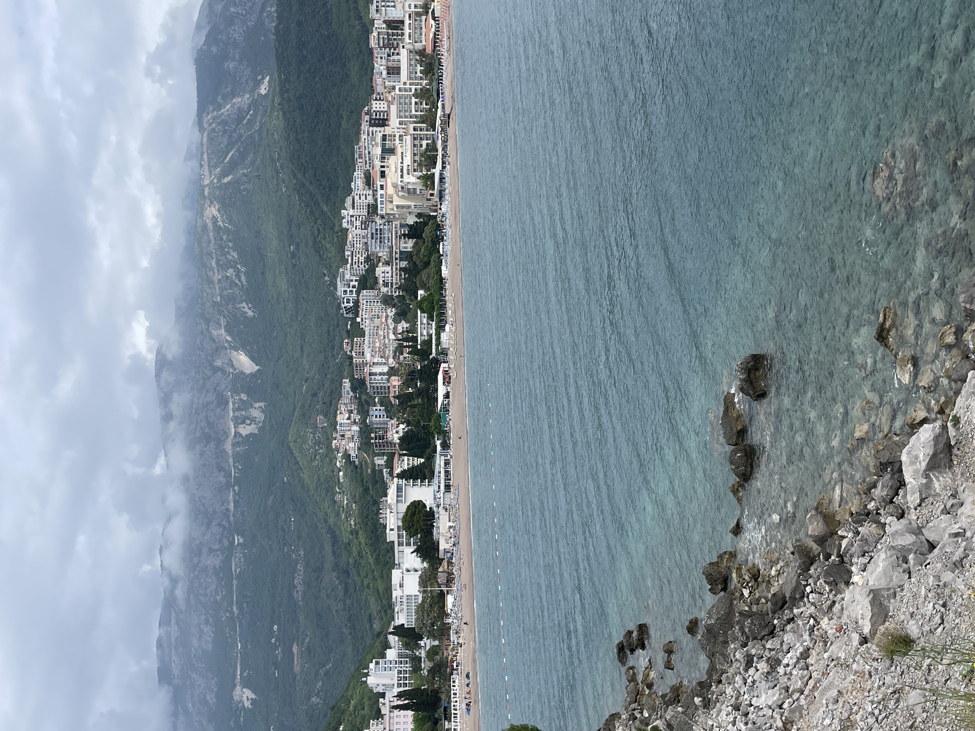 The image shows a coastal view in Budva, Montenegro, featuring a long sandy beach lined with hotels and buildings at the base of a steep, forested mountain shrouded in low-hanging clouds. The clear turquoise water of the Adriatic Sea stretches out to the right, with visible rocks along the immediate shoreline. The beach is marked with swimming area buoys and dotted with people and sun loungers. The perspective is from an elevated position, possibly a cliffside.