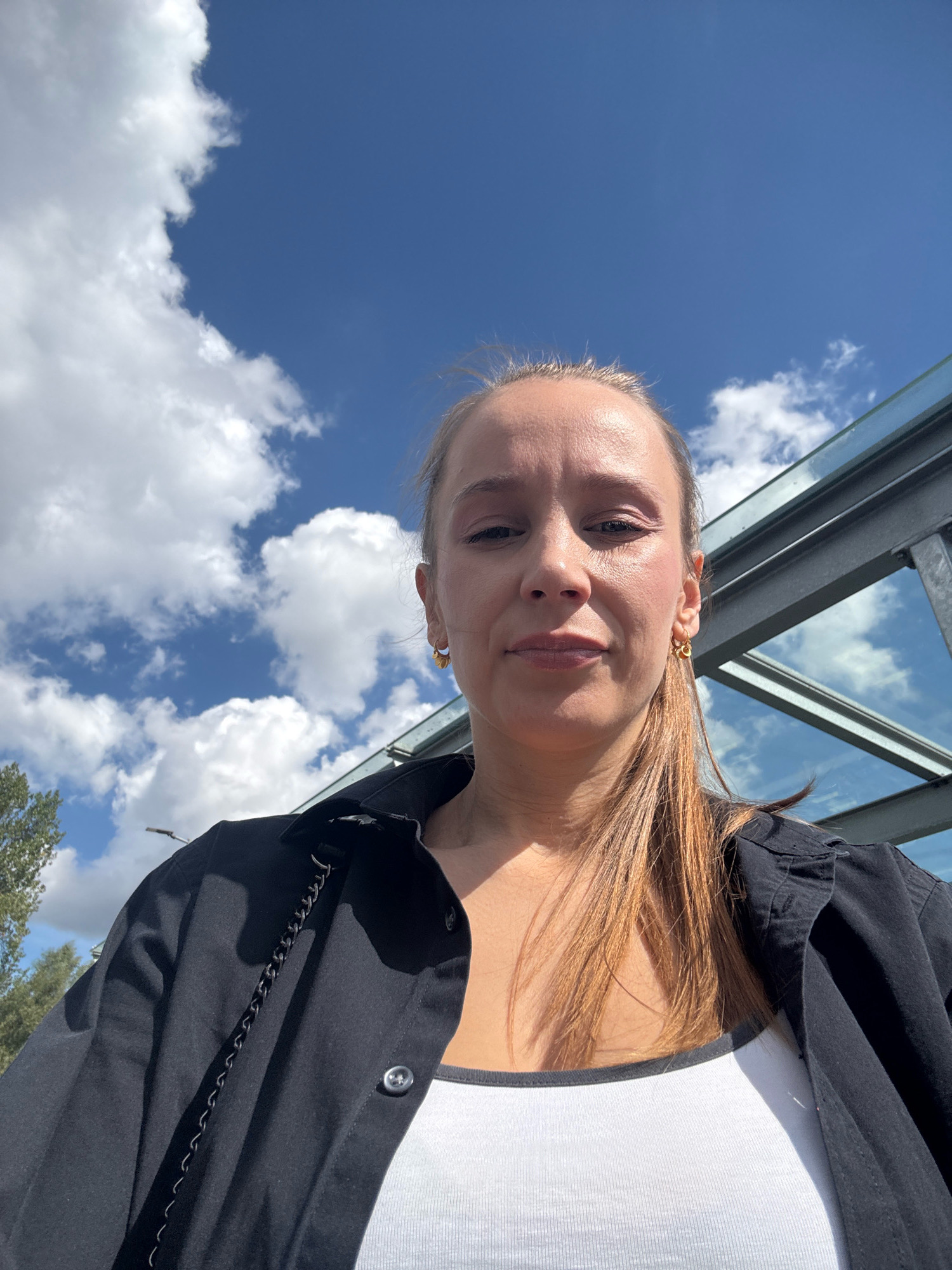 A low-angle, full-body selfie of a woman with blonde hair pulled back in a ponytail, wearing a black button-up shirt over a white tank top and gold hoop earrings. She is looking directly at the camera with a neutral expression. The background shows a bright blue sky with scattered white clouds and a glimpse of a modern glass structure, possibly a walkway or building canopy. Trees are visible on the left side of the image. The scene appears to be outdoors, possibly in a public area. The location is specified as Überherrn, Germany.