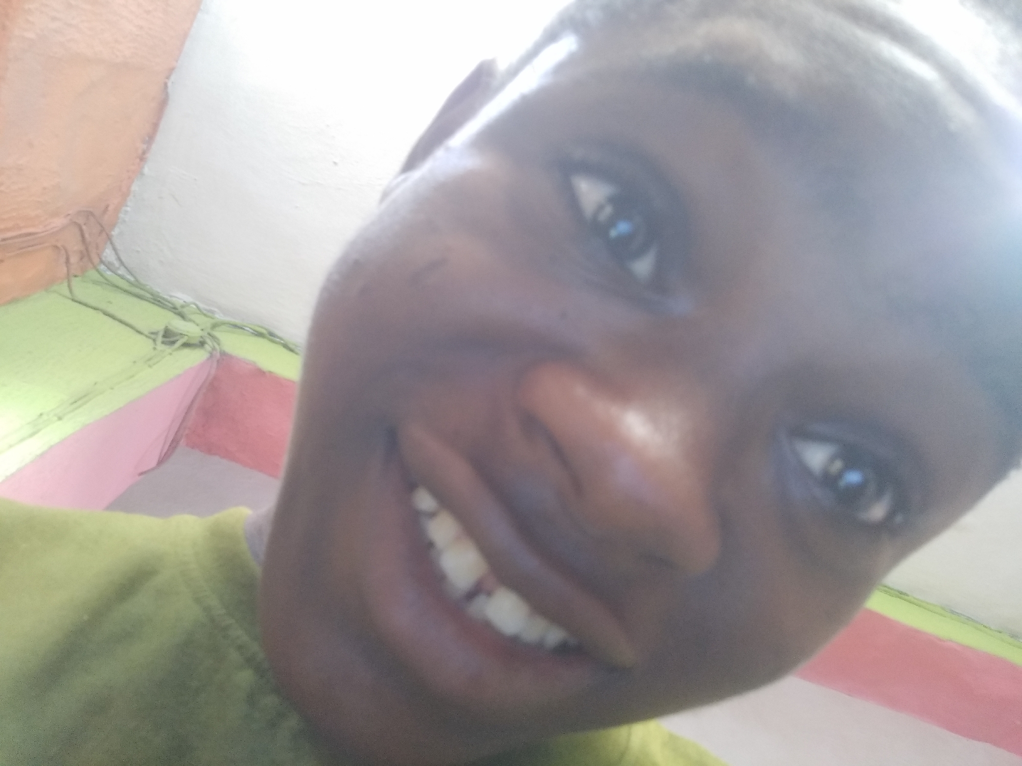 A close-up, low-angle selfie of a dark-skinned person with short hair, smiling and looking slightly upward. The person is wearing a green shirt. In the background, a wall is visible, painted with horizontal bands of white, pink, and green. Electrical wires run along the top of the wall where it meets the ceiling. The location is likely Shangisha, Nigeria, based on the request.