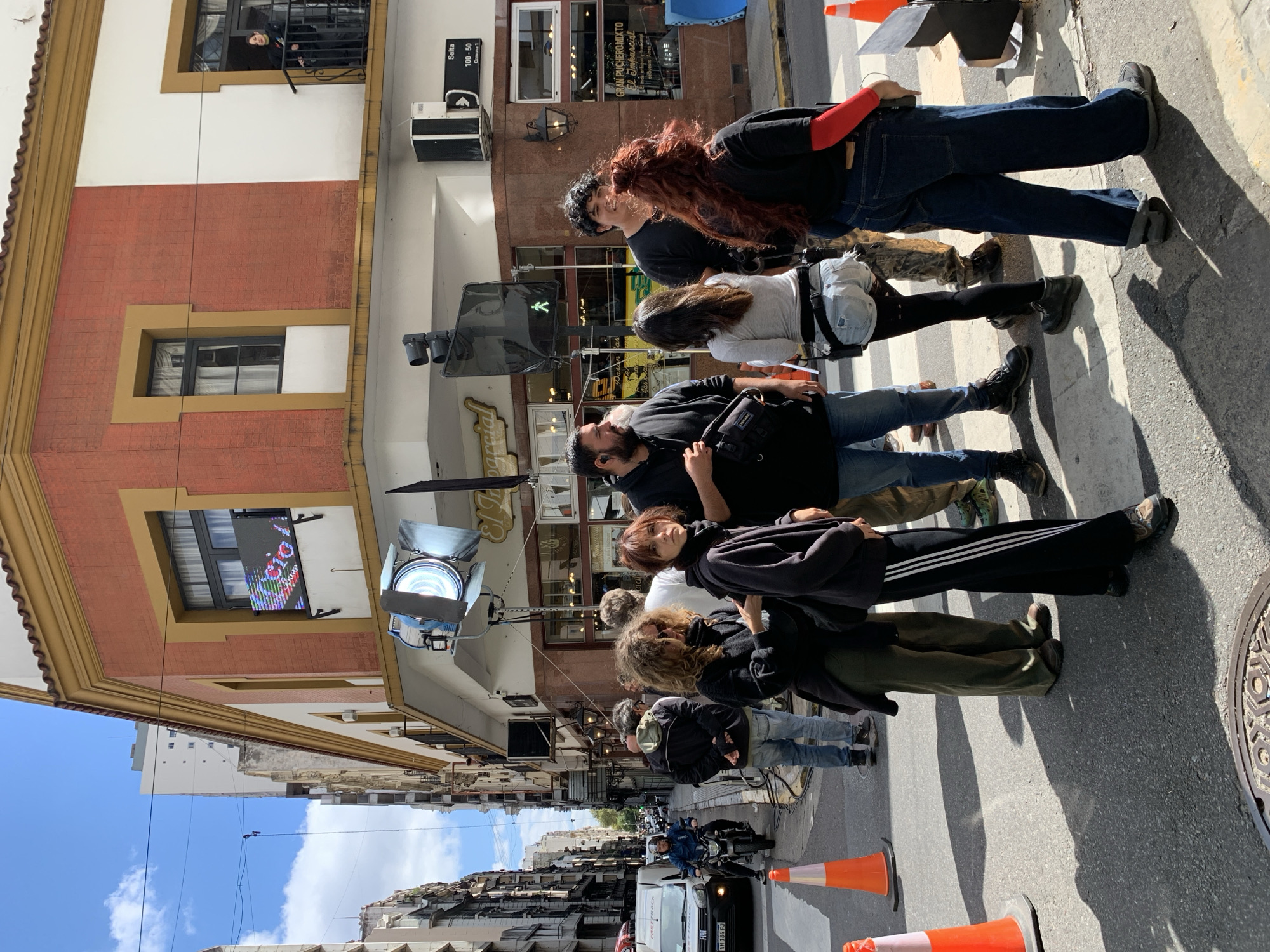 The image captures a scene in Buenos Aires, Argentina, specifically on a street corner where a film or video shoot appears to be in progress.
Several people are standing or walking on a zebra crossing, some seemingly part of the production crew and others likely pedestrians. The crowd includes individuals with diverse clothing styles, from casual wear to more distinctive outfits.
Filming equipment, including a large spotlight, is visible on the side of the street, indicating the presence of a film crew.
The background features buildings with distinctive architectural details. A sign "Salta 100-50 Comuna 1" is visible, suggesting a location in the area of Salta street in the Comuna 1 district of Buenos Aires. In addition, the storefront signs such as "El Comercial" are visible. A person is seen standing on a balcony of one of the buildings.
Traffic cones mark off the area, controlling traffic flow and pedestrian access. A van marked with "Fast Track" is on the street, as is a scooter with a person on it. The overall impression is that of a bustling street where a film shoot is temporarily disrupting normal activity.